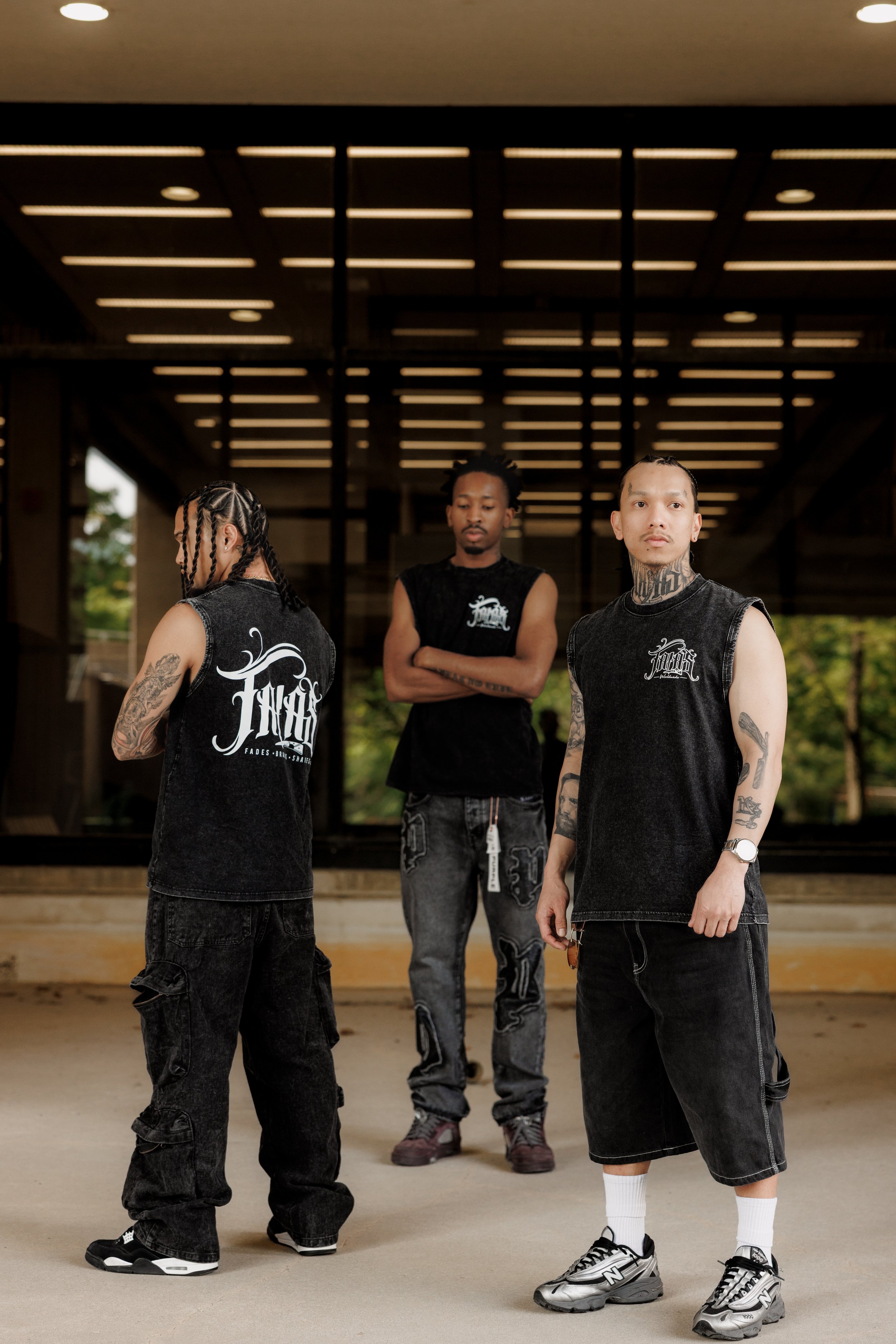 Three young men with tattoos and wearing black sleeveless shirts with white logos pose outdoors under a wooden pergola; they are wearing black cargo pants and sneakers, one is looking directly at the camera, while the others are looking away or with 