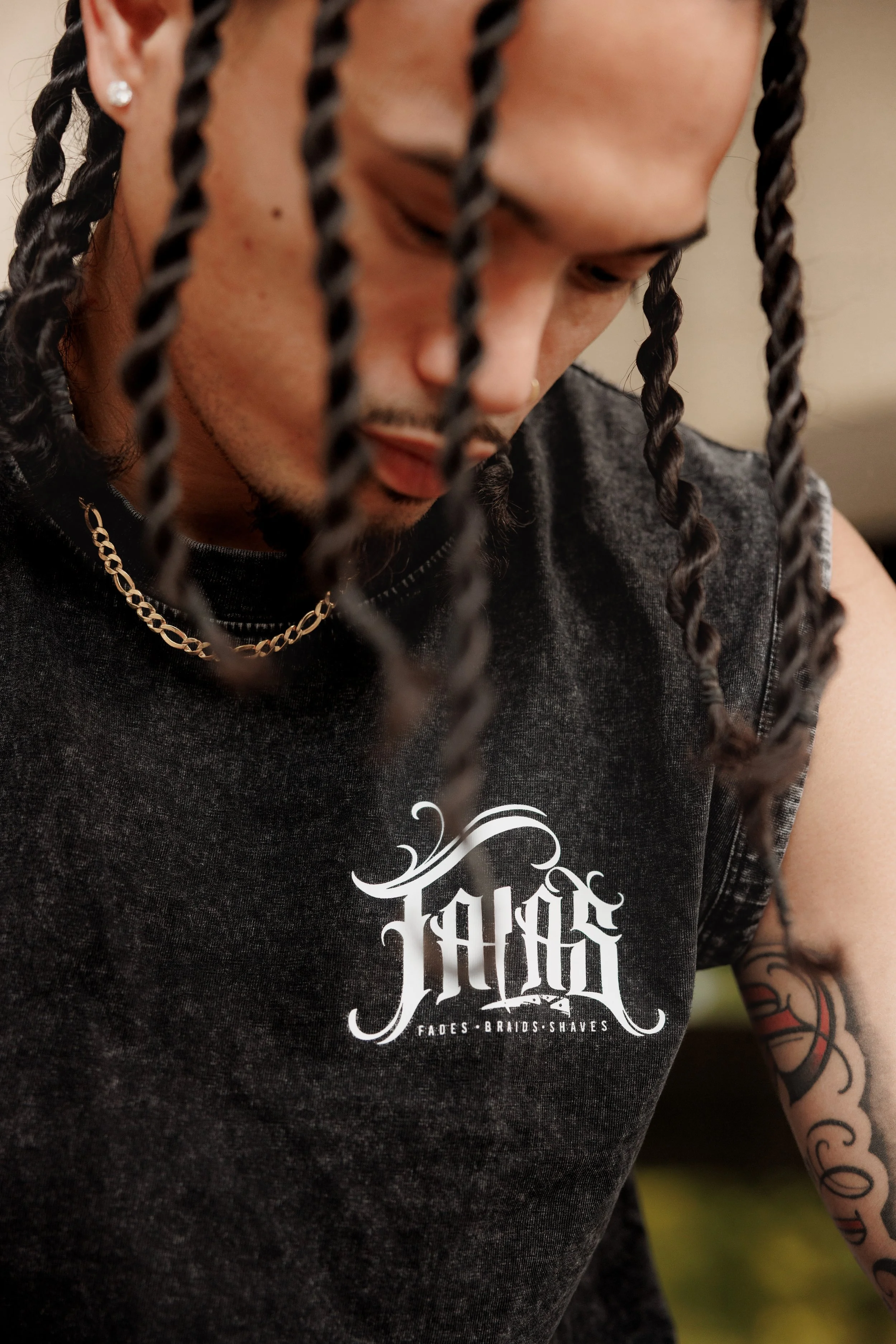 A man with braided hair, wearing earrings and a gold chain, is looking down. He is wearing a dark T-shirt with white gothic-style text that reads 'Fades, Braids, Shaves'.