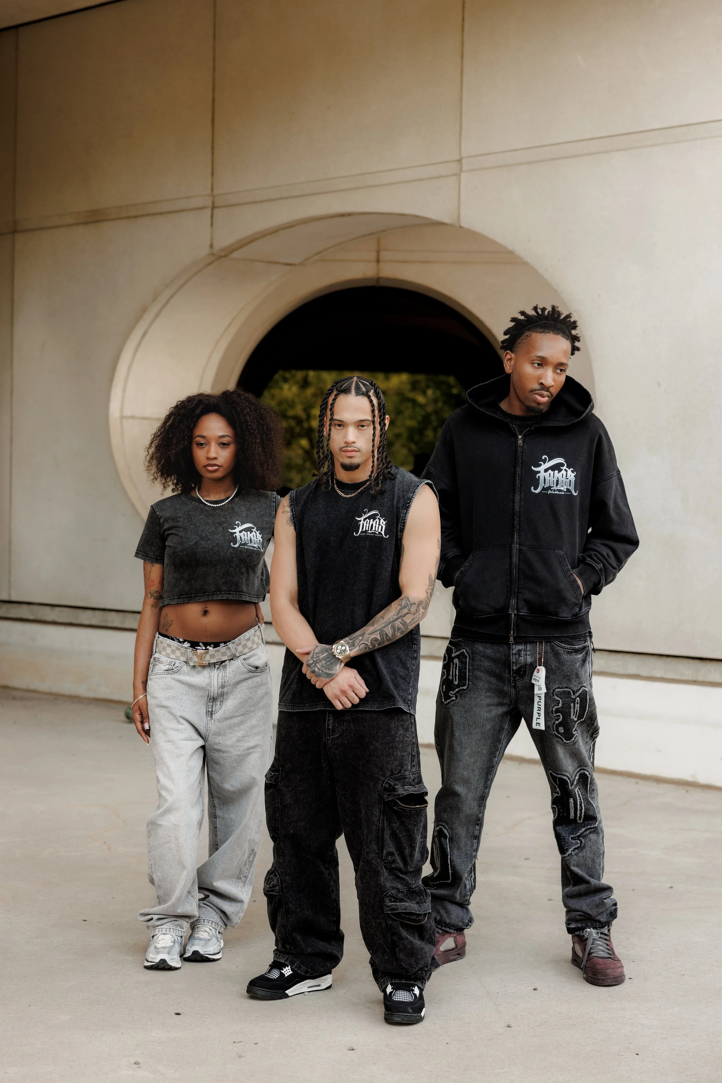 Three young adults walking outside in front of a concrete archway, wearing casual streetwear and sneakers.