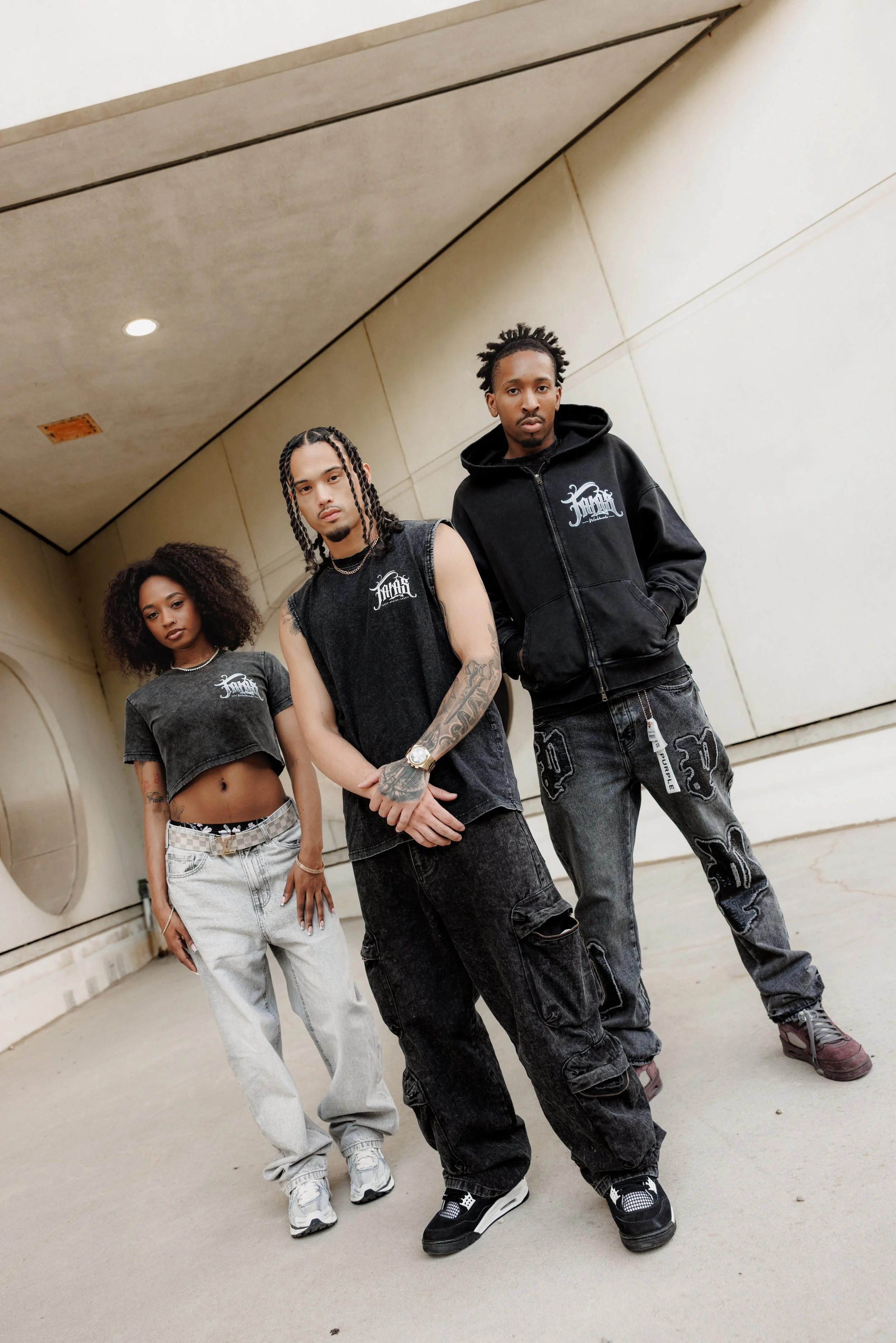 Three young adults standing in a covered outdoor area with beige walls and ceiling. They are dressed in streetwear, with two men and one woman, all with serious expressions.