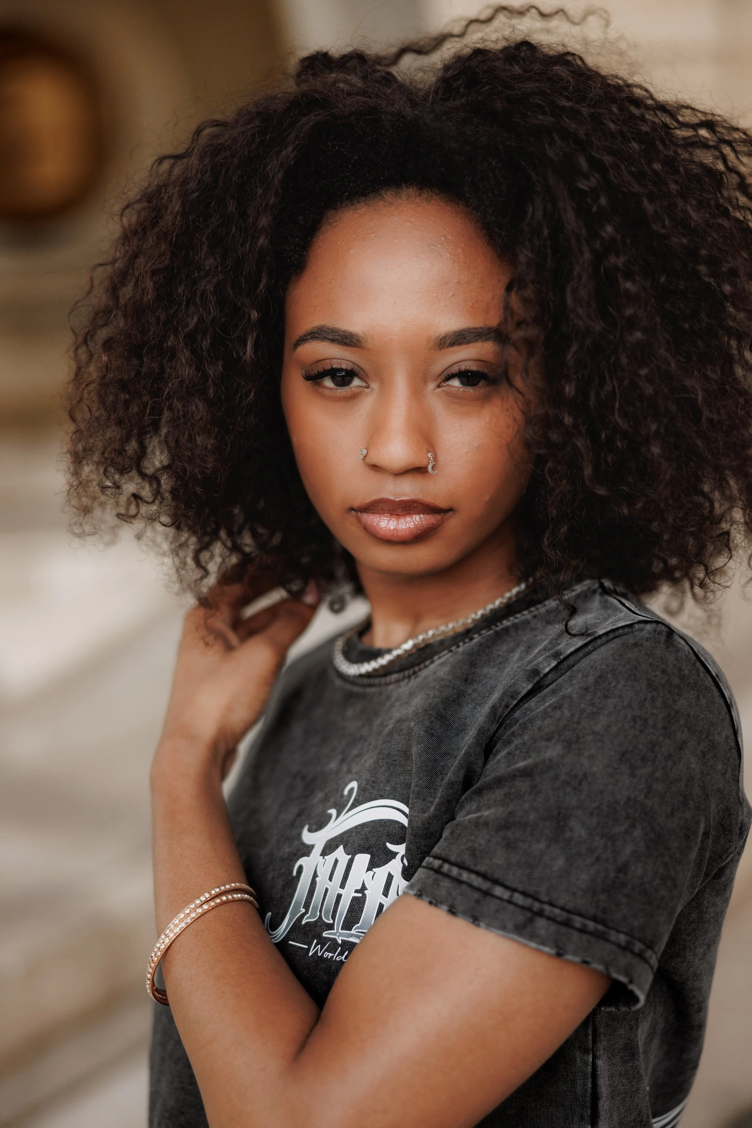 A woman with dark curly hair and nose piercings wearing a black t-shirt with white text, accessorized with bracelets and a necklace, standing outdoors.
