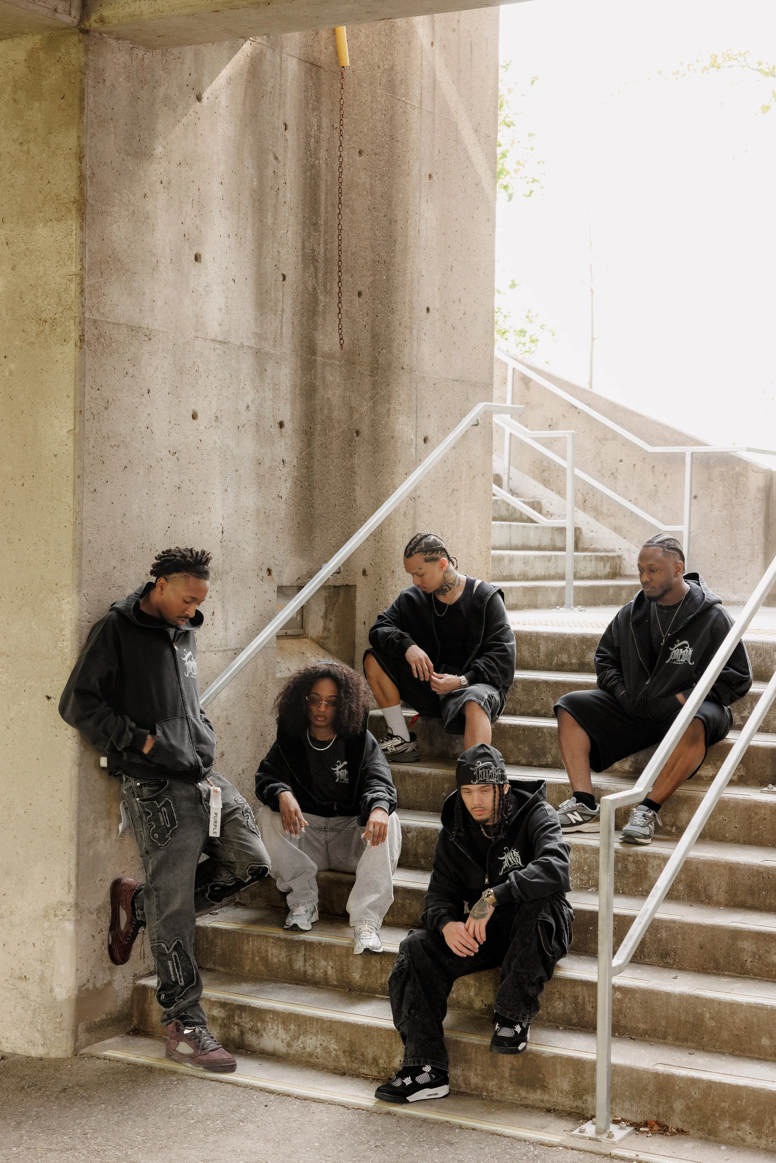 Group of six young people sitting and standing on concrete stairs outdoors, with a concrete wall and railing, dressed in casual streetwear, some wearing hoodies and sneakers, in a relaxed pose.