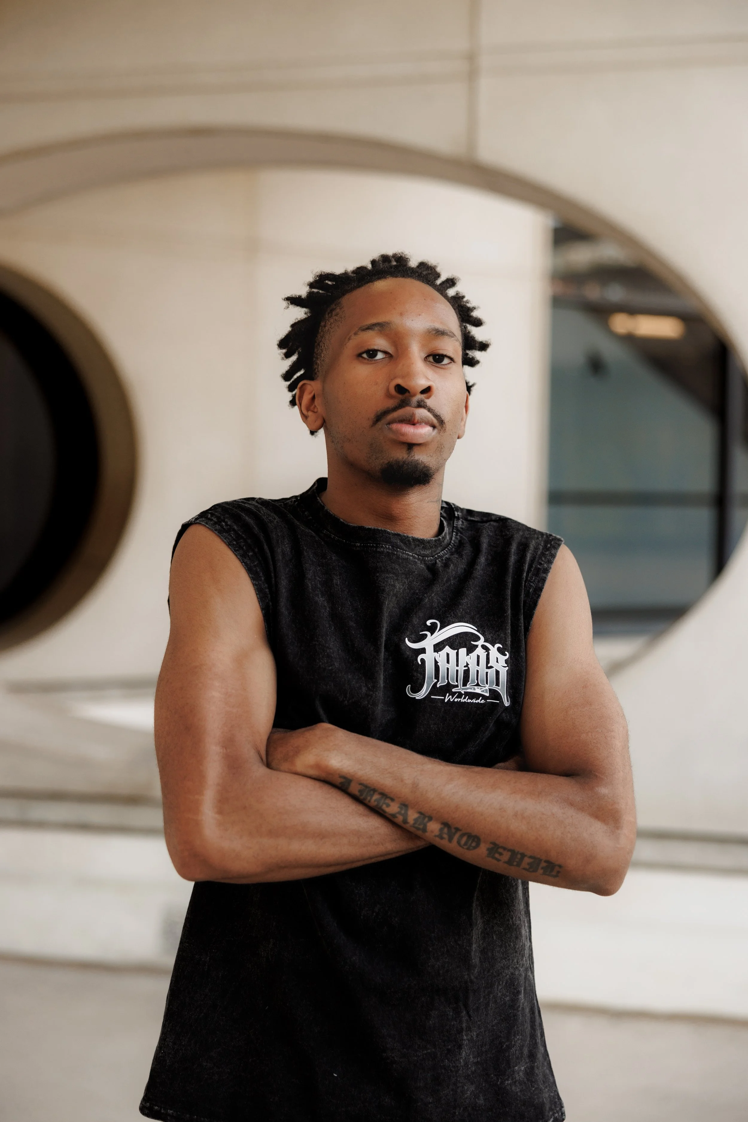 A young man with dreadlocks and a mustache, wearing a sleeveless black shirt, standing with arms crossed in front of a circular mirror in a modern indoor space.