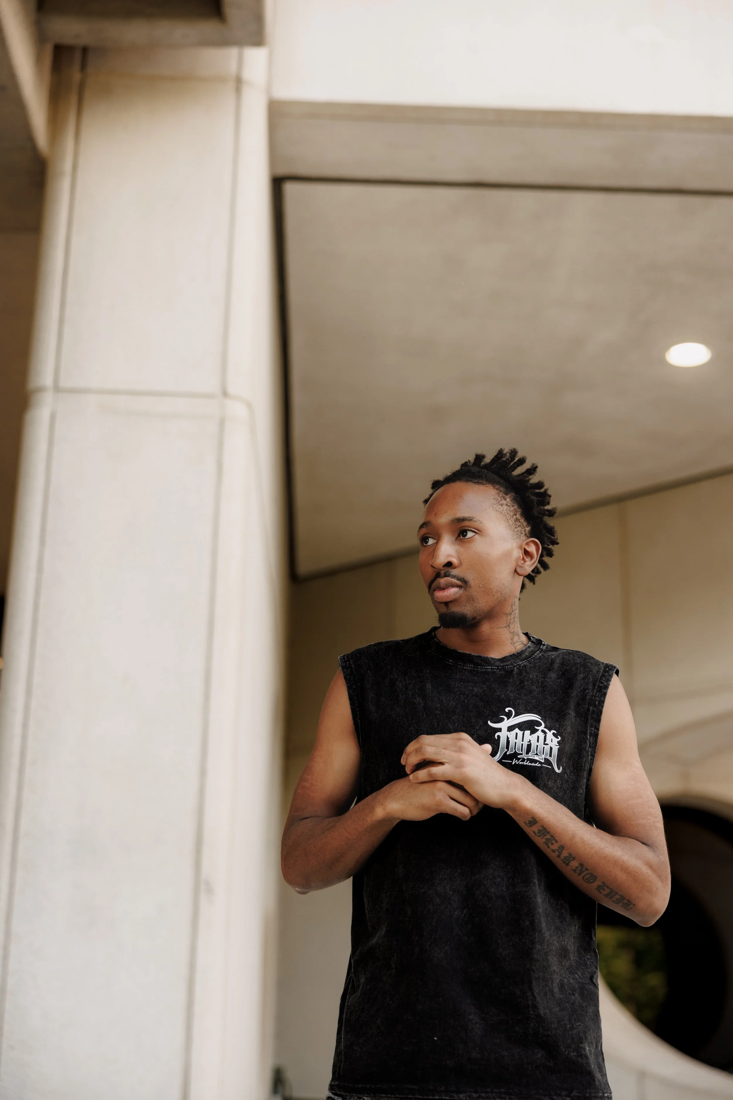 A young man with dreadlocks and tattoos on his arms standing indoors, wearing a black sleeveless shirt with white text on it.