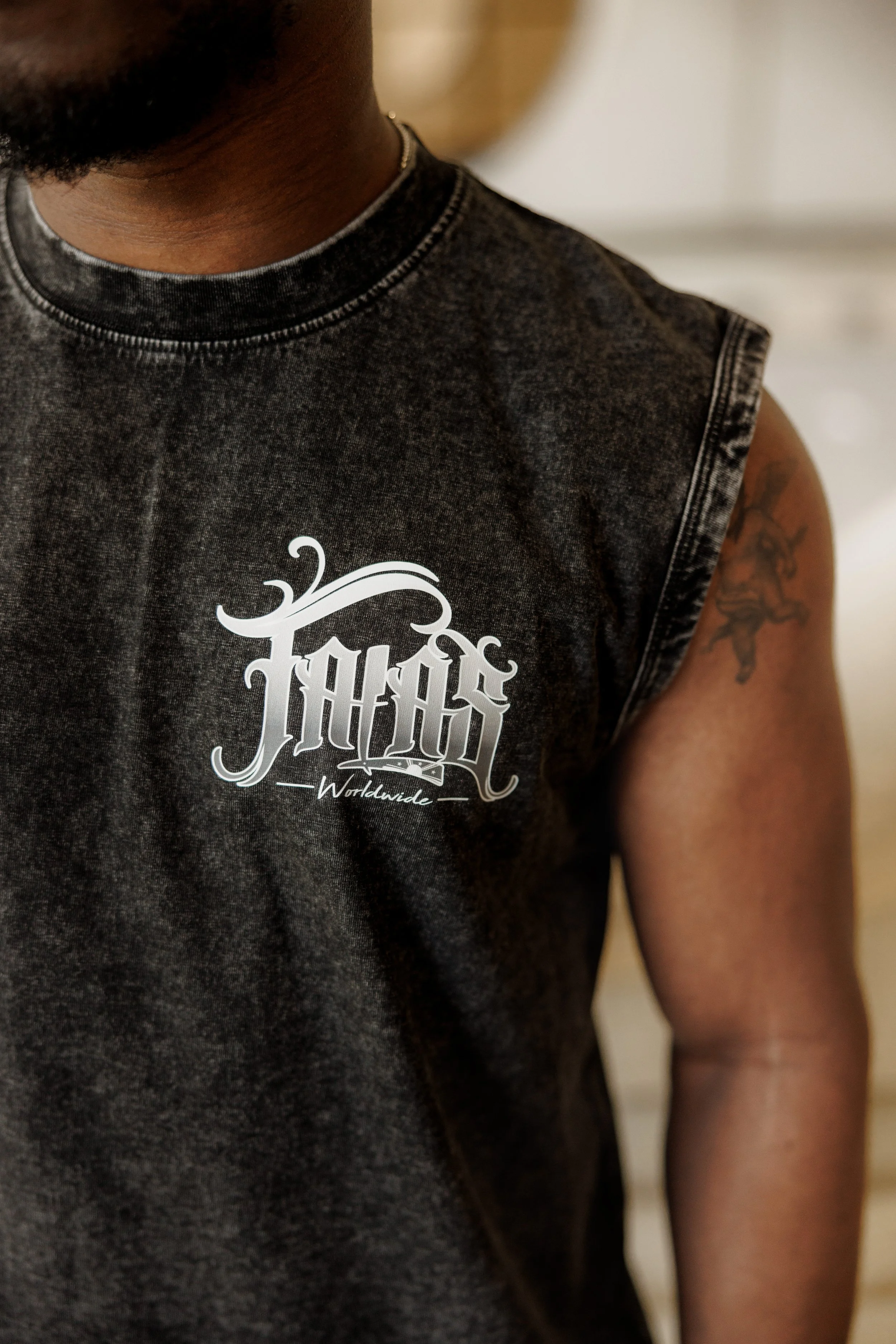 Close-up of a person wearing a sleeveless, faded black T-shirt with a white graphic design that reads 'TFO' and 'Worldwide'. The person has dark skin, a beard, and a tattoo on their upper arm.