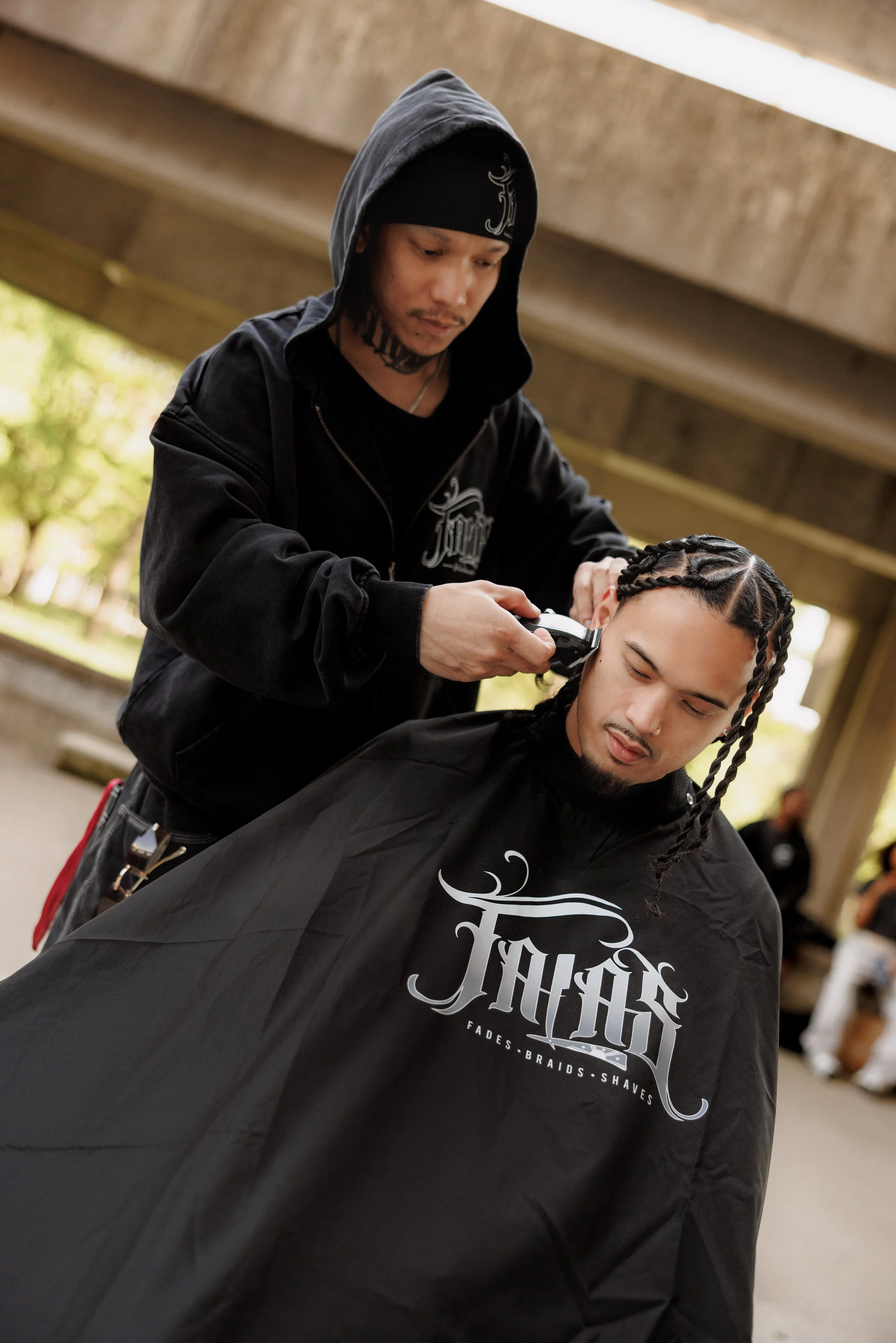 A person is getting a haircut outdoors under a bridge. The barber is wearing a black hoodie and beanie, and is using clippers on a client's hair. The client has braided hair and is wearing a black cape with white text.