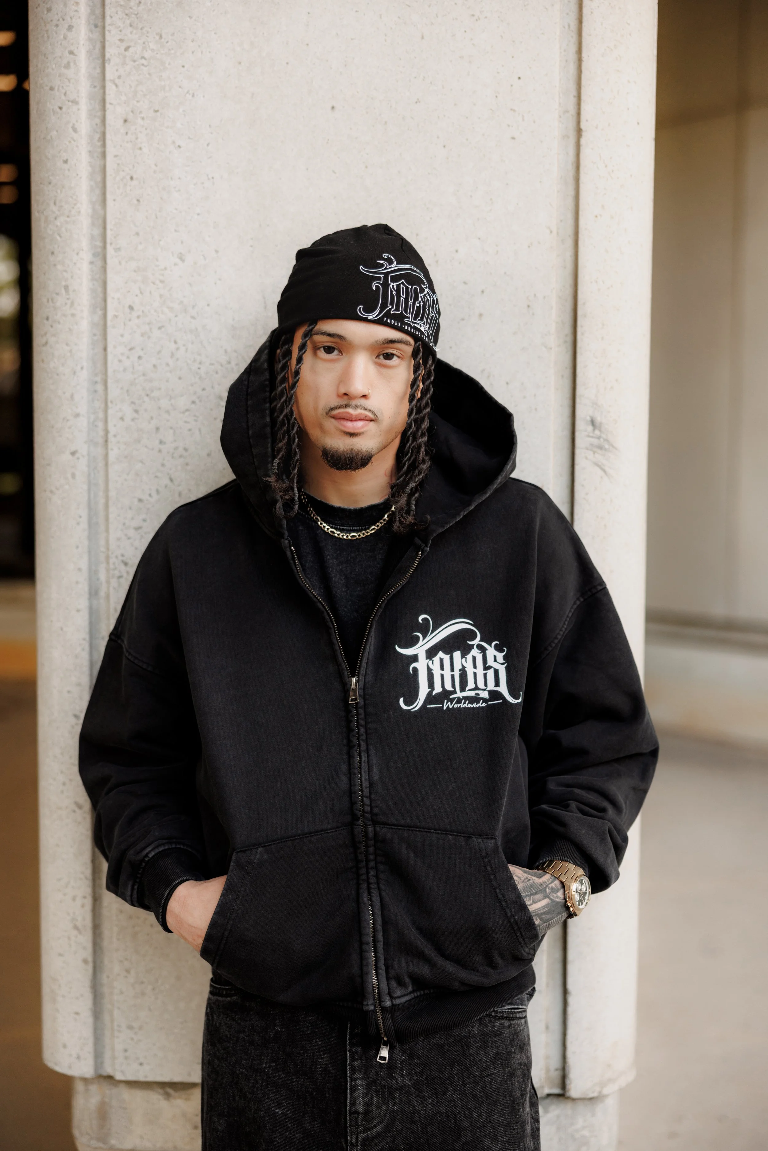 A young man with long braided hair, wearing a black beanie with white text, black hoodie with a white logo, dark jeans, gold watch, and chains, standing against a light-colored stone wall.