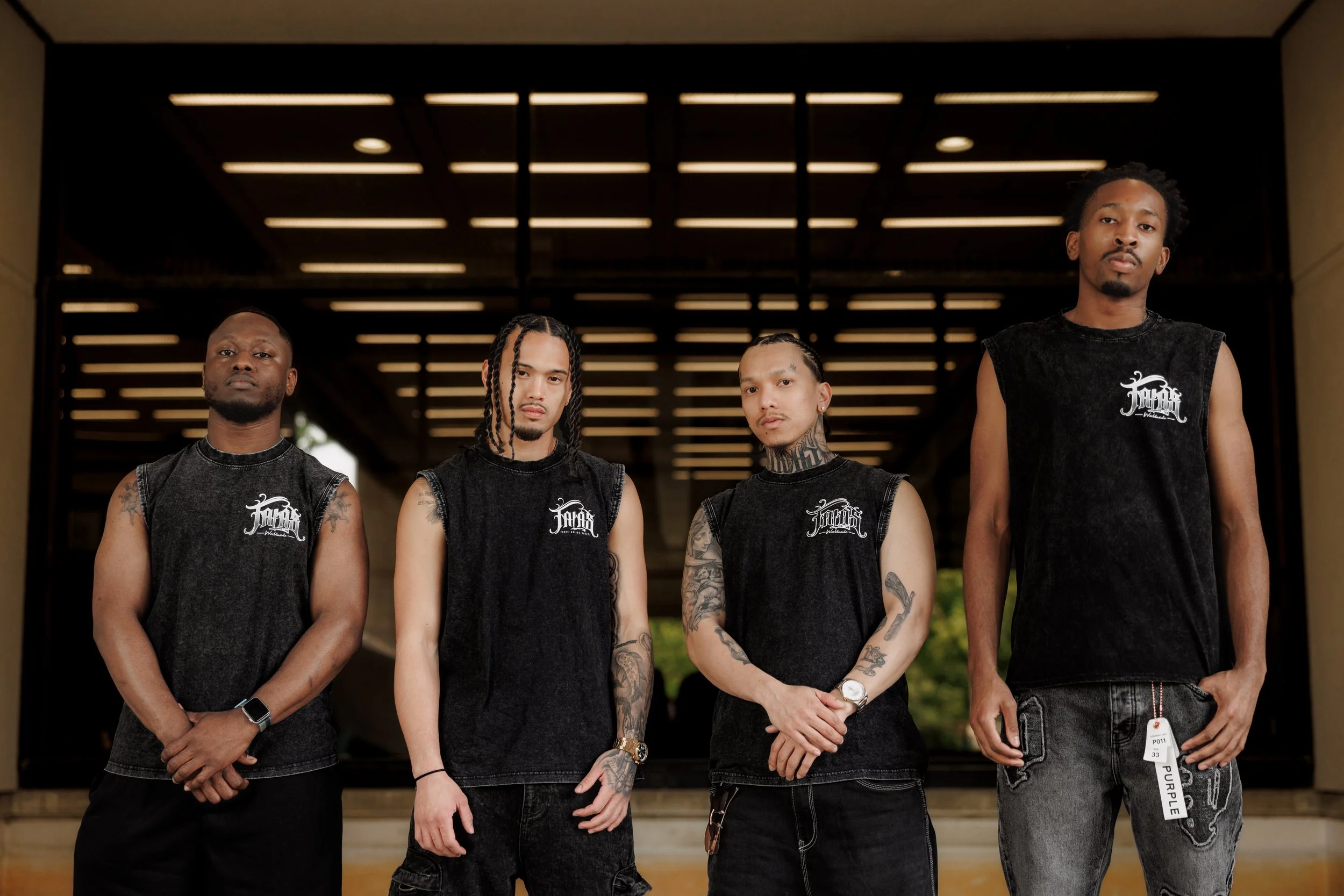 Four young men standing outdoors under an architectural structure with horizontal lighting. They are wearing sleeveless black shirts with white logo and have tattoos, with serious expressions.