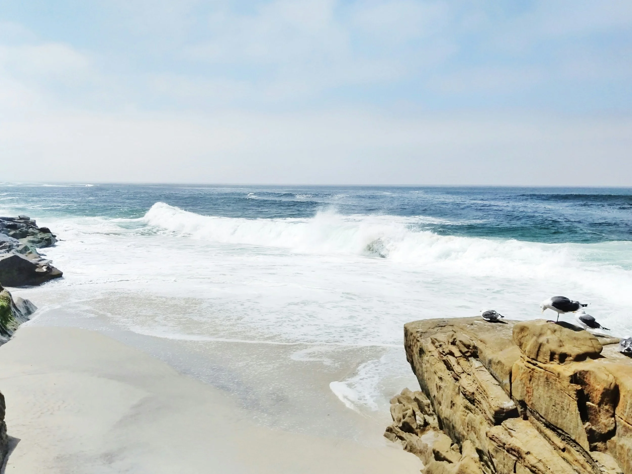 A beach scene with ocean waves crashing against rocks and sandy shore, with a partly cloudy sky overhead.