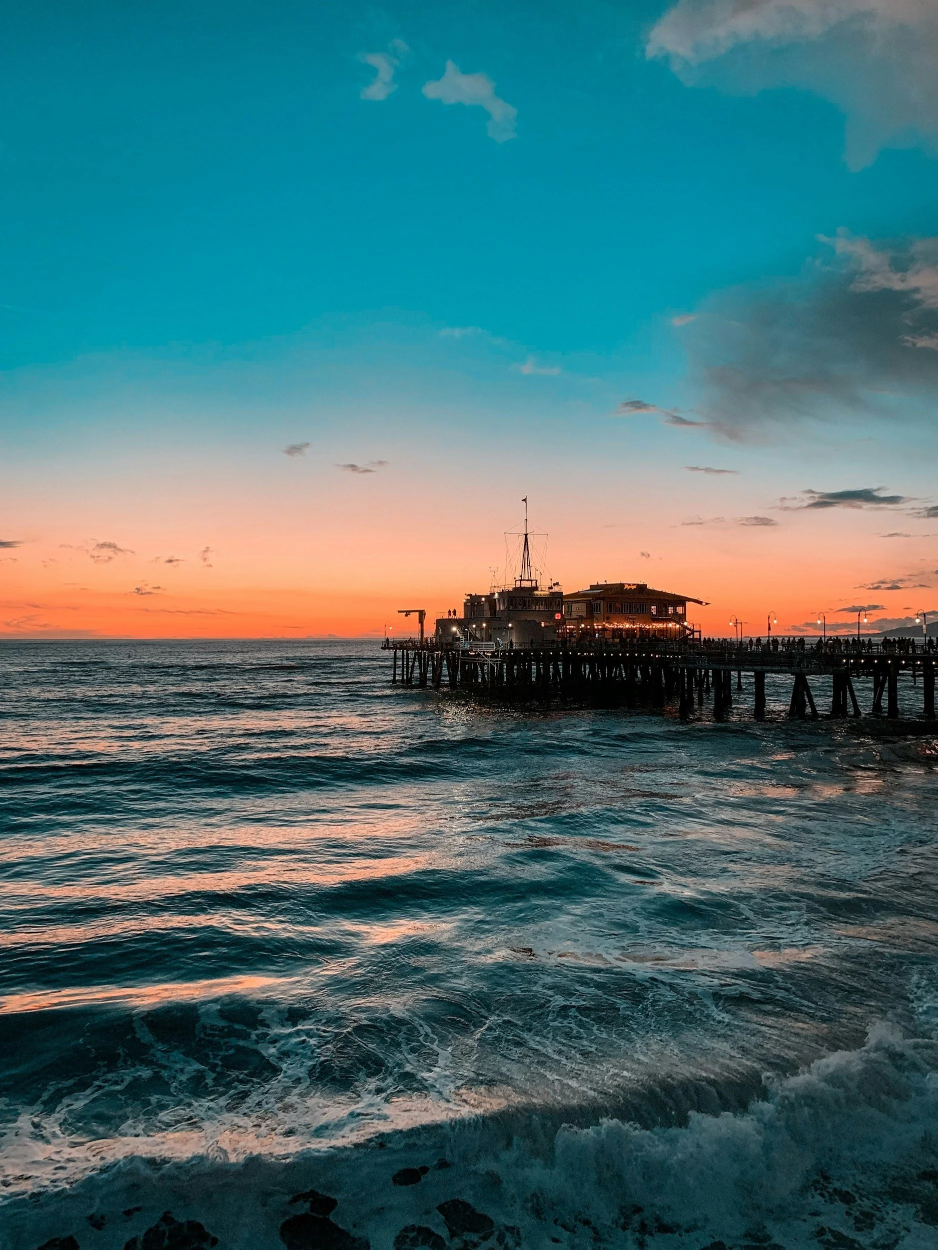 Sunset over the ocean with a pier extending into the water and buildings at the end, with a sky transitioning from orange to blue