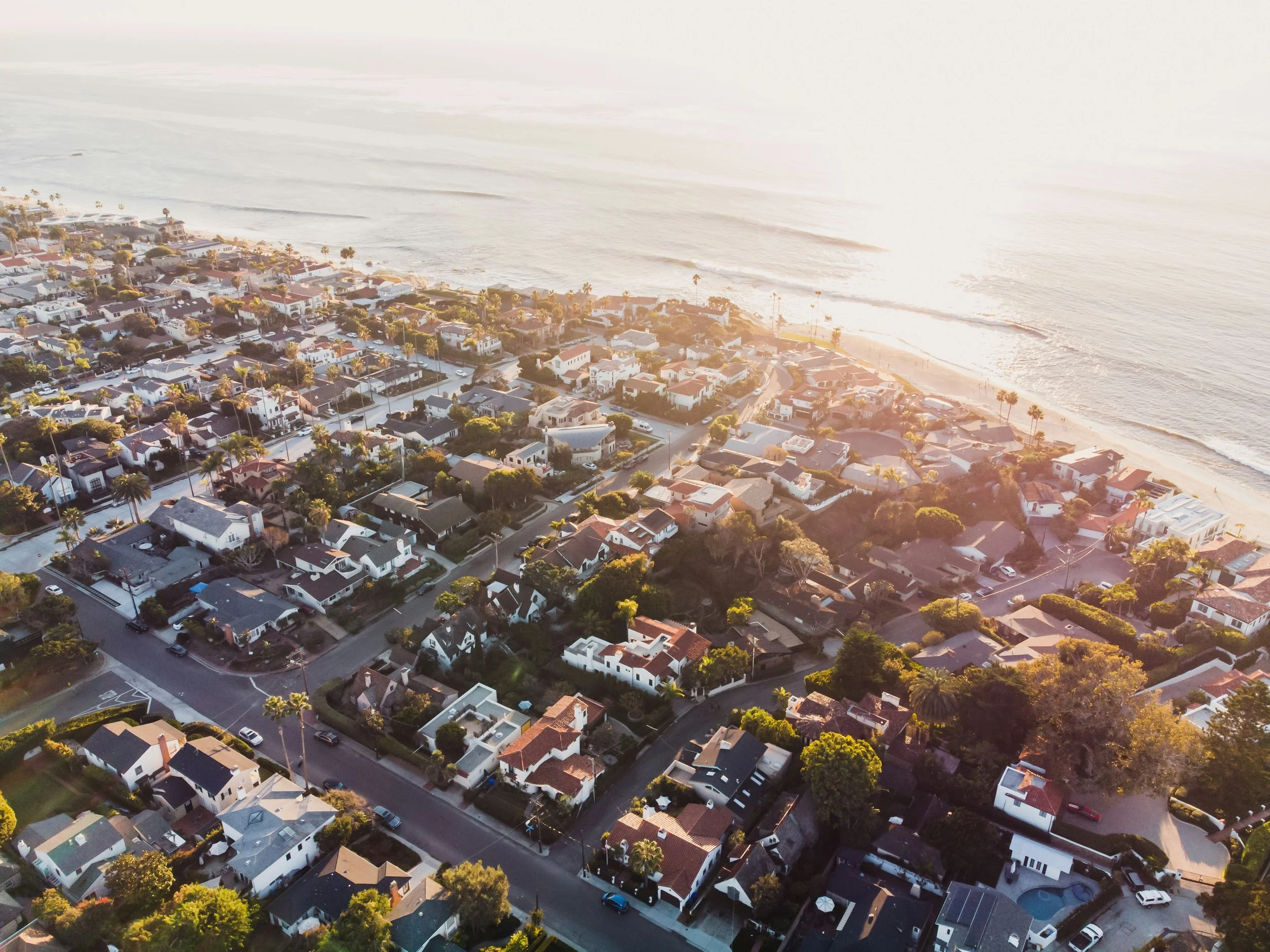 An aerial view of a coastal neighborhood with houses, palm trees, and streets adjacent to the beach and ocean during sunset.