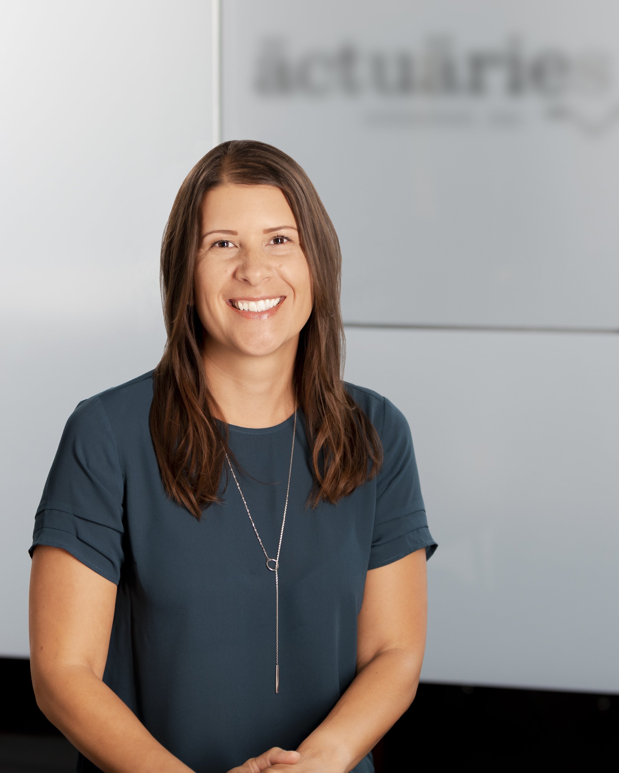 Smiling woman with brown hair wearing a dark teal top standing in front of a blurred office background with a sign that appears to read 'AboutMe'.