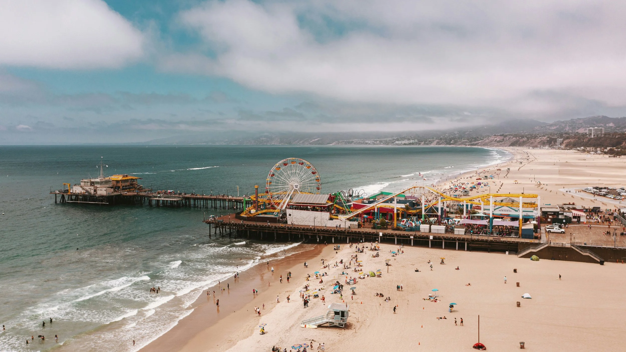 A beach with a pier, amusement park rides, and many people sunbathing and walking on the sand, under cloudy skies.