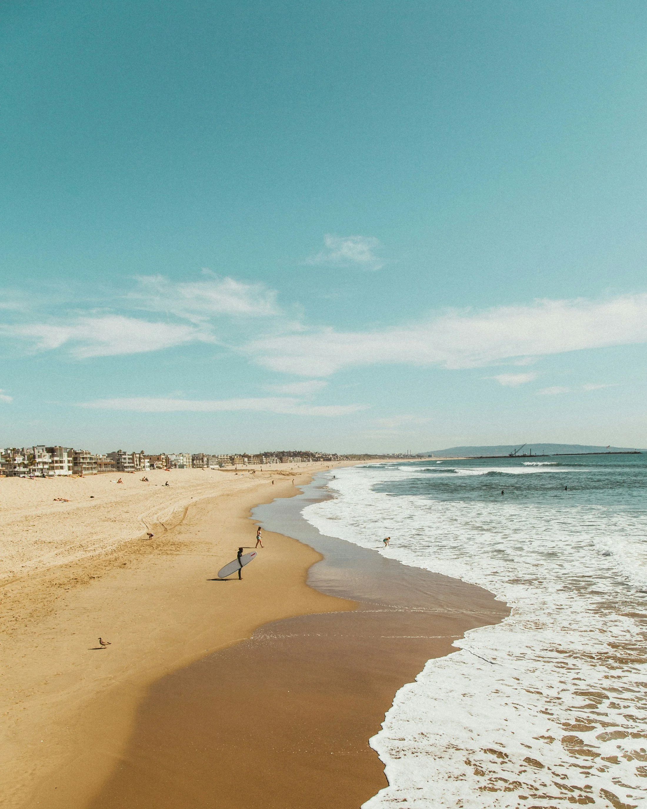 A beach scene with sand, ocean waves, and a clear blue sky. There are a few people enjoying the beach, some with surfboards, and beachfront buildings in the background.