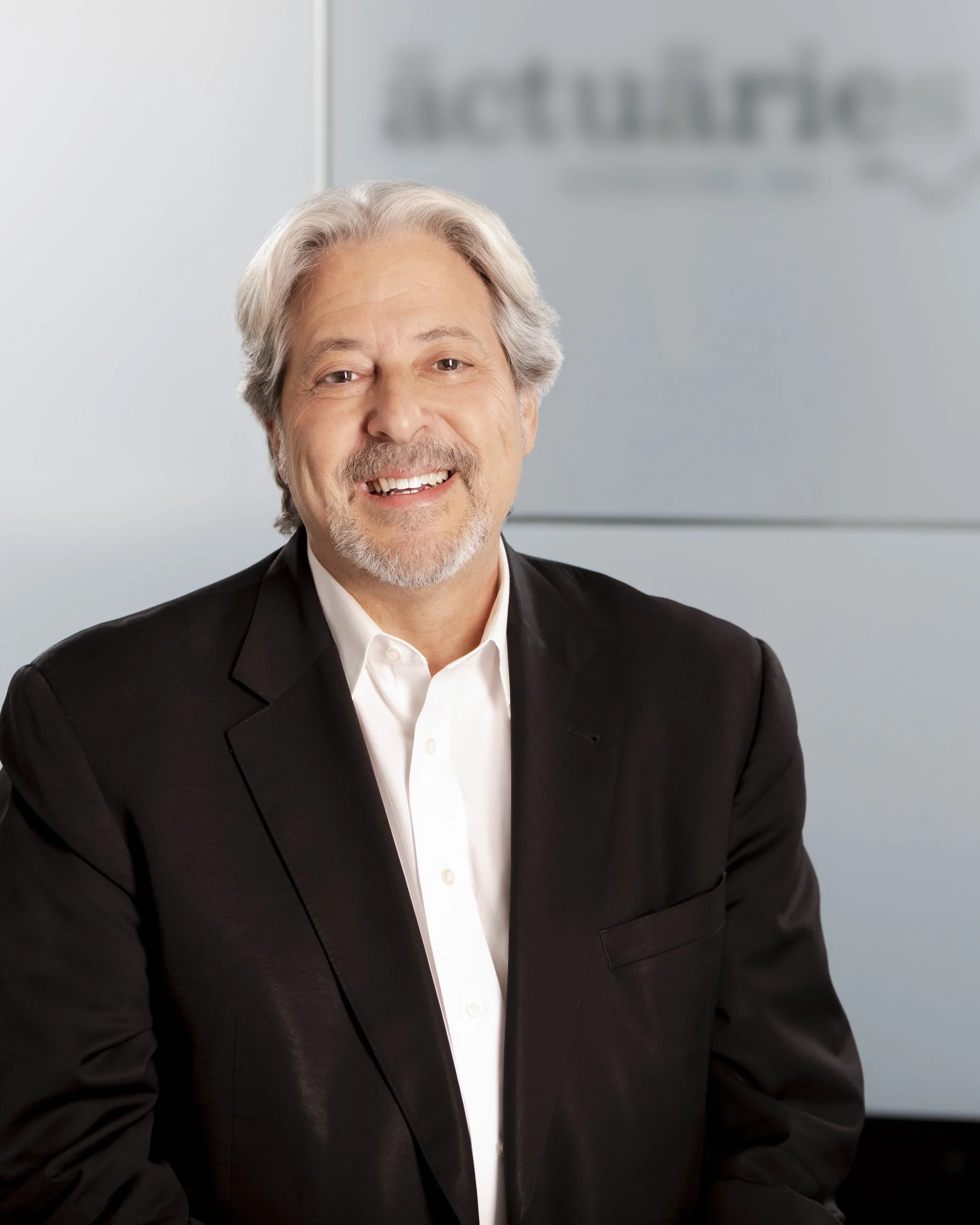 Close-up of a smiling man with gray hair and a beard, wearing a black suit and white shirt, with a blurred background featuring a sign that appears to say 'Auditorium.'