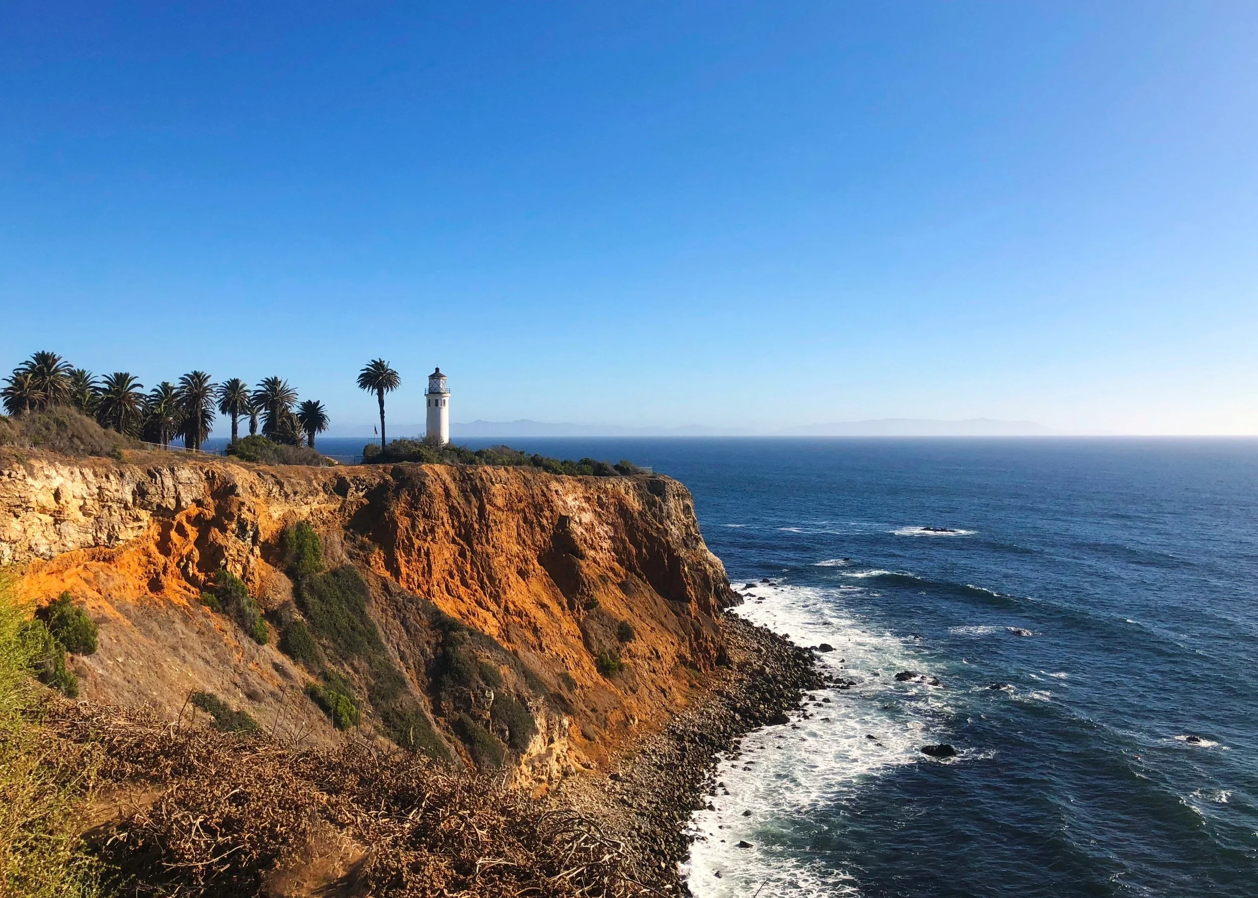 Coastal landscape with cliffs, a lighthouse, palm trees, and the ocean under a clear blue sky.
