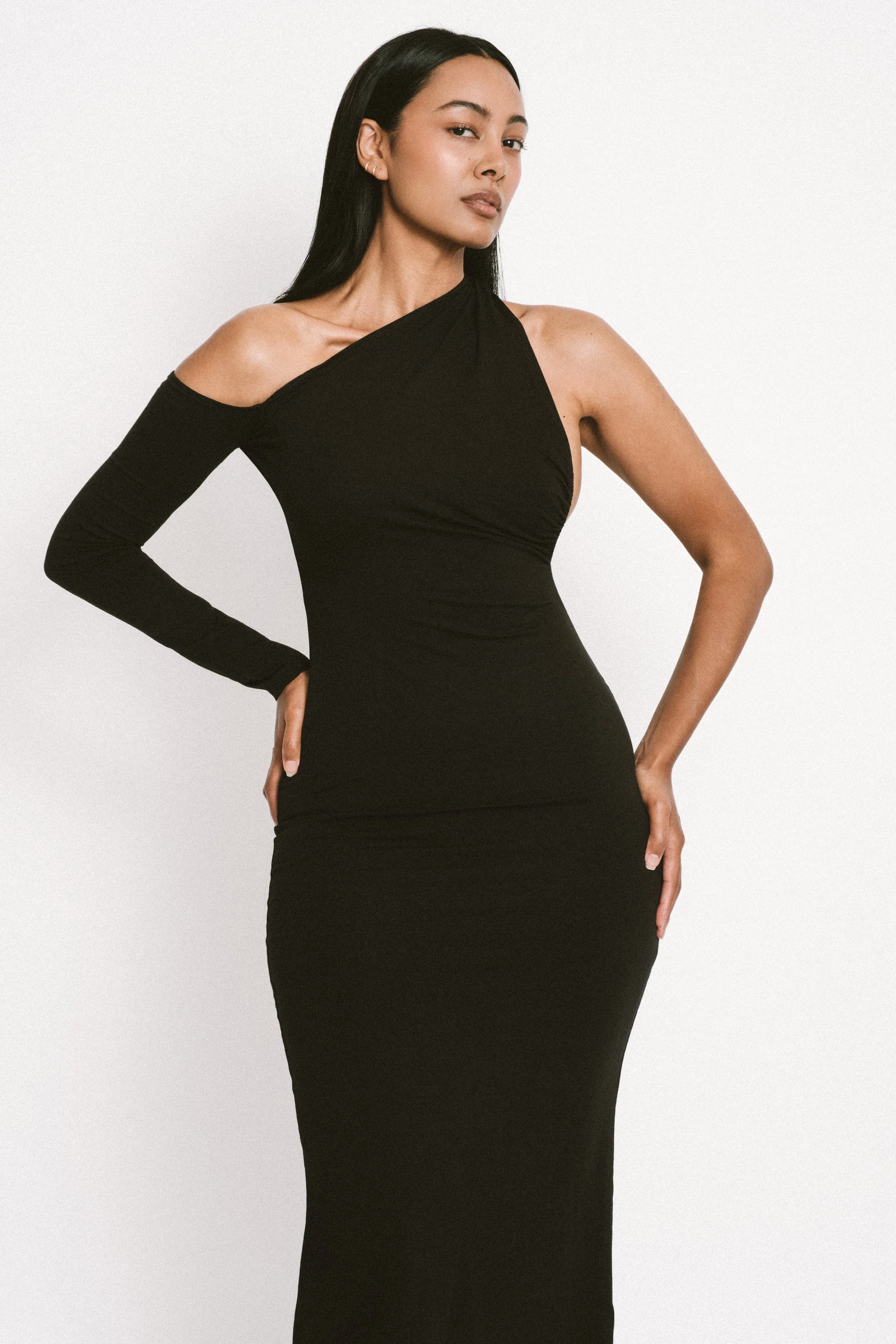 Luxury lifestyle portrait of Jennifer Elysee wearing a minimalist black one-shoulder asymmetric evening dress against a neutral studio backdrop.