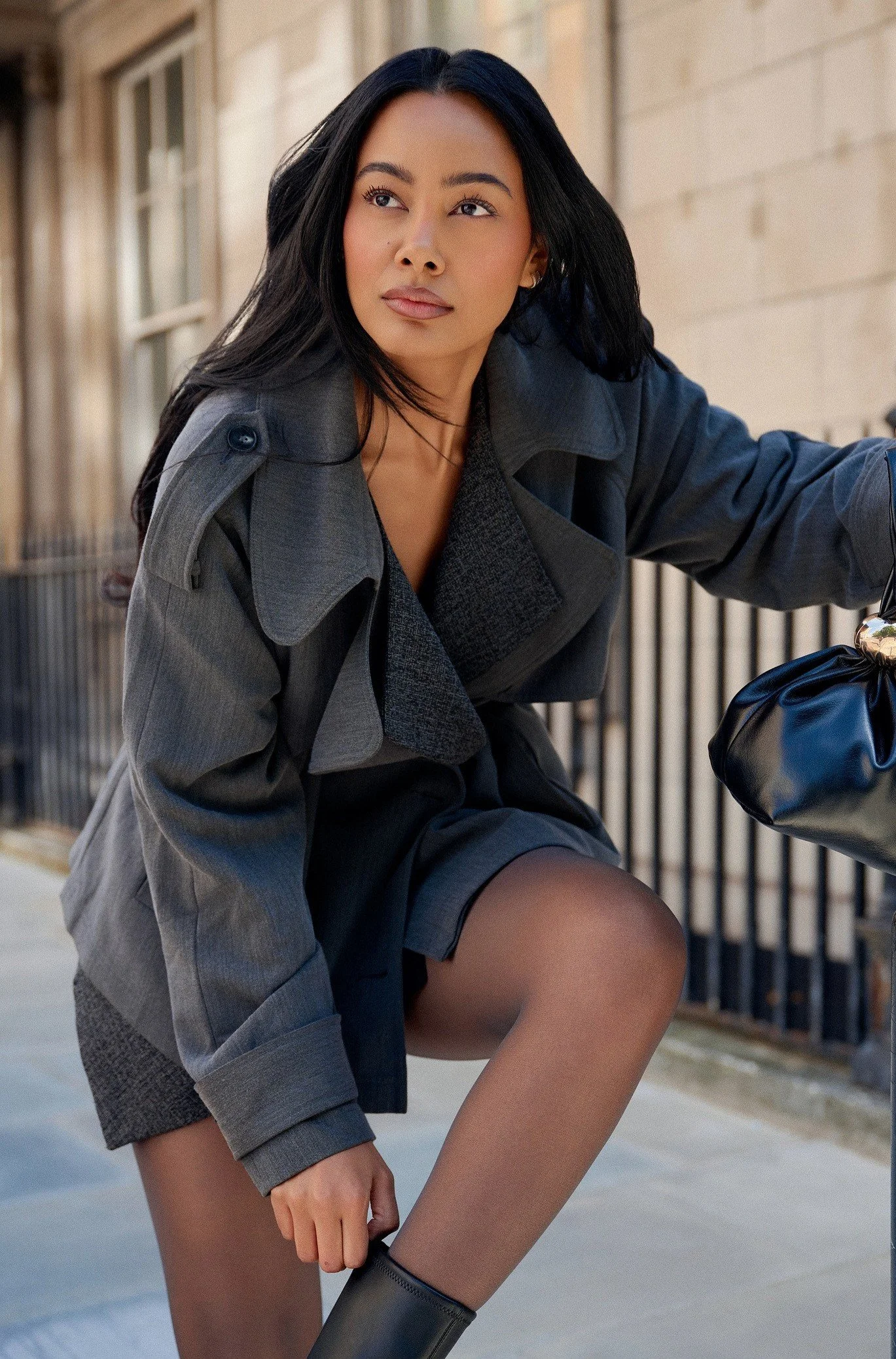 Editorial fashion portrait of Jennifer Elysee crouching in a grey trench coat with black ankle boots and a designer handbag.