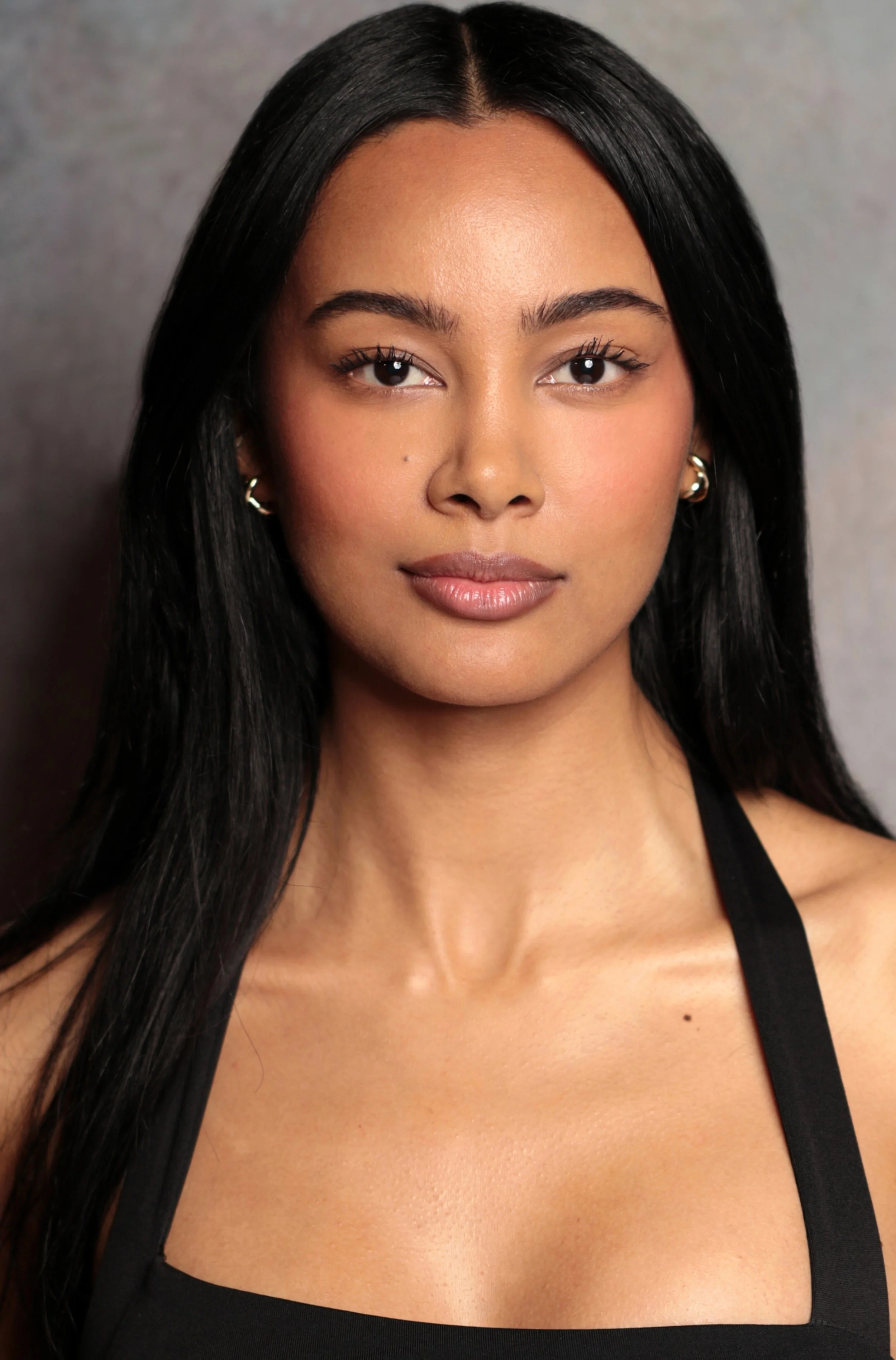 Close-up beauty portrait of Jennifer Elysee with natural makeup and sleek dark hair against a dark background.