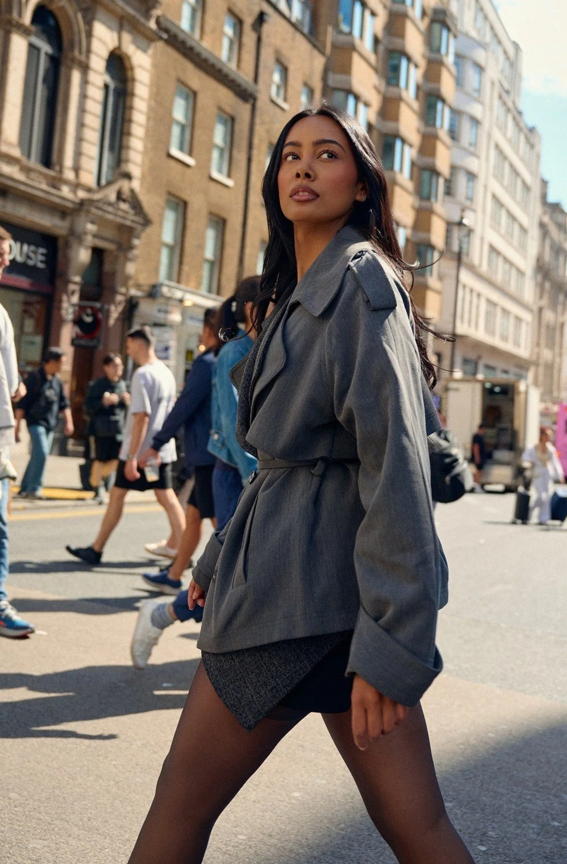Luxury fashion campaign portrait of Jennifer Elysee walking through a London street wearing a tailored grey trench coat.