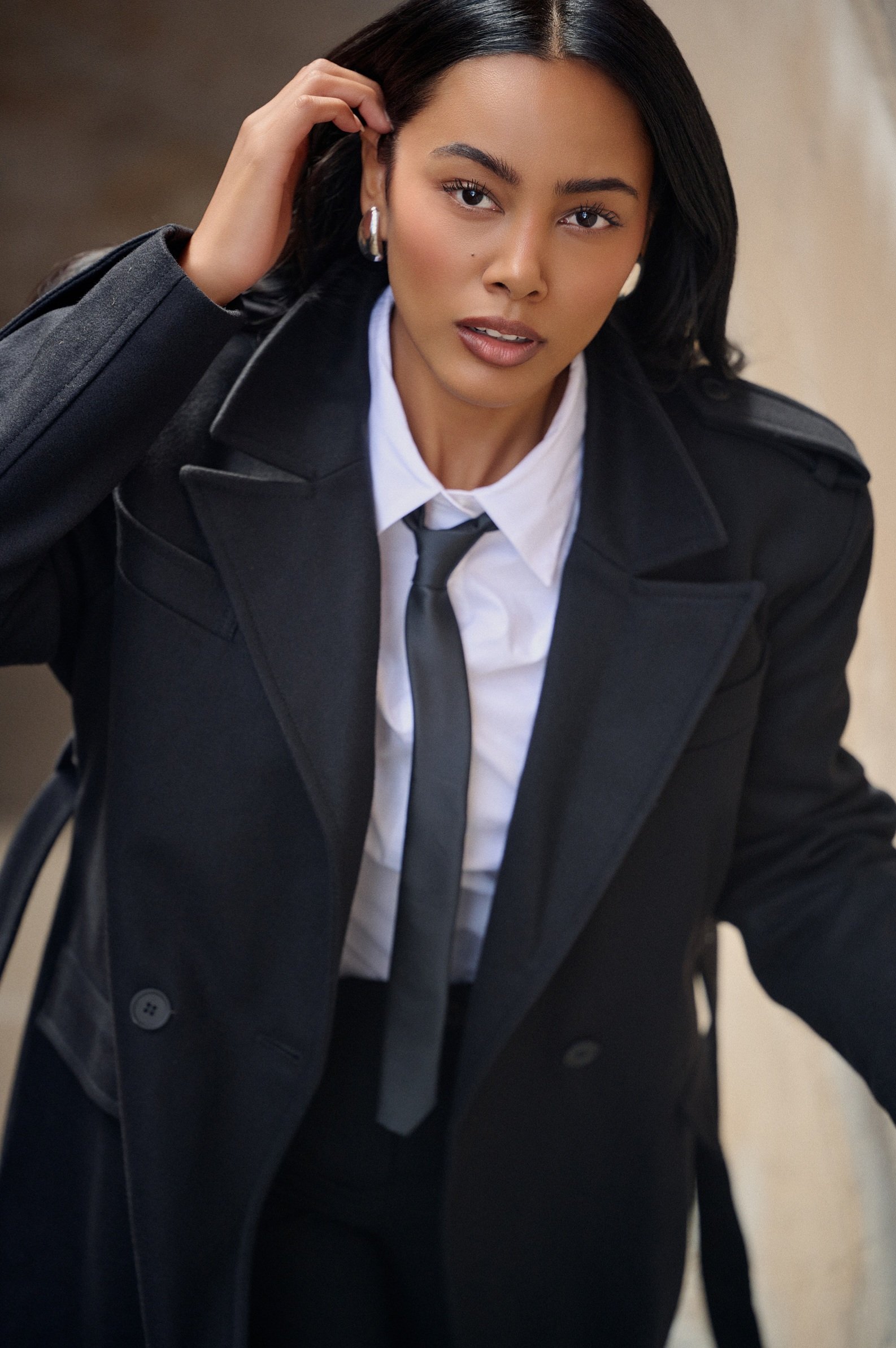Editorial fashion portrait of Jennifer Elysee wearing a black coat layered over a white shirt and tie in a luxury city setting.