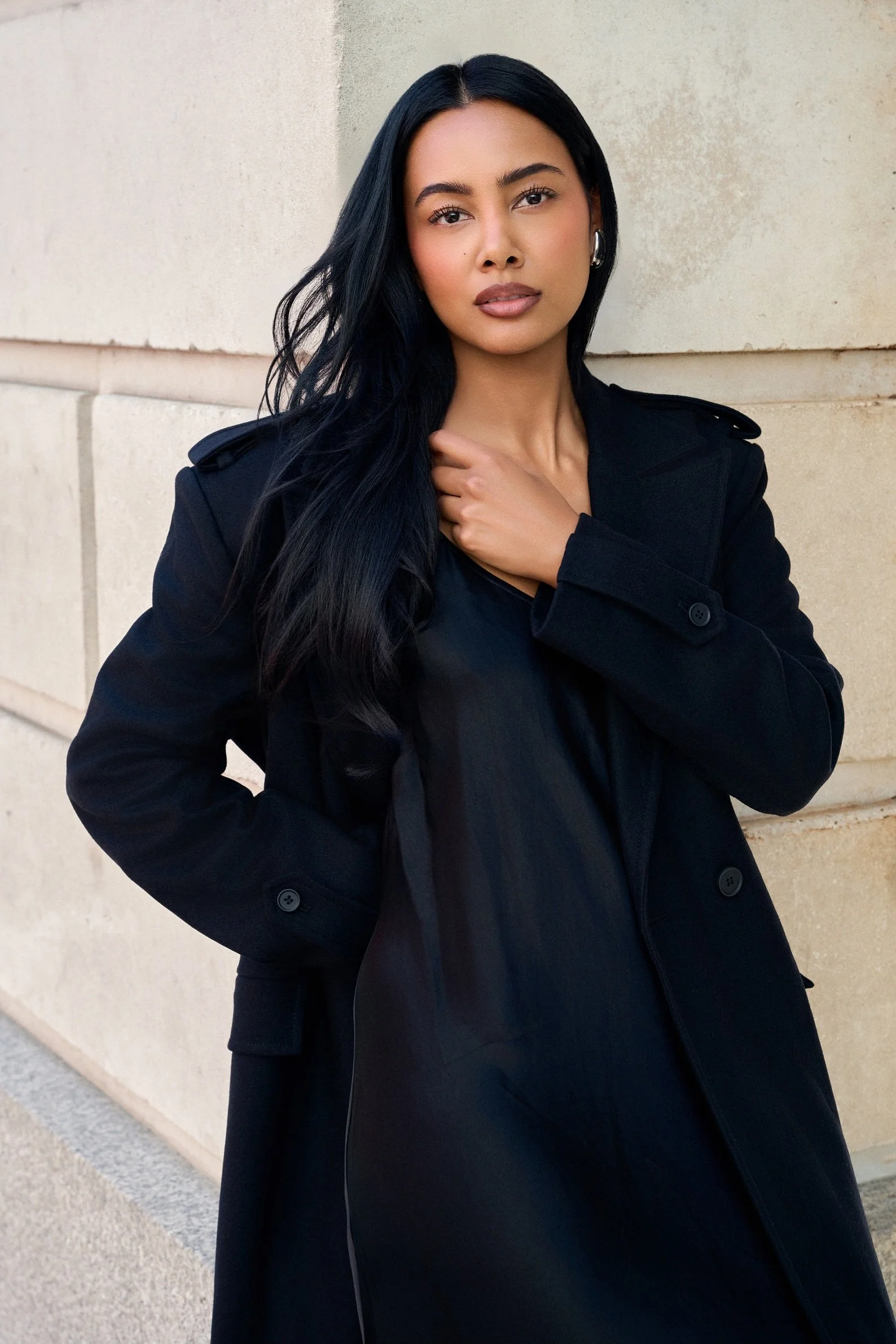 Luxury fashion portrait of Jennifer Elysee wearing a black tailored coat with sleek hair and natural makeup against a stone wall.