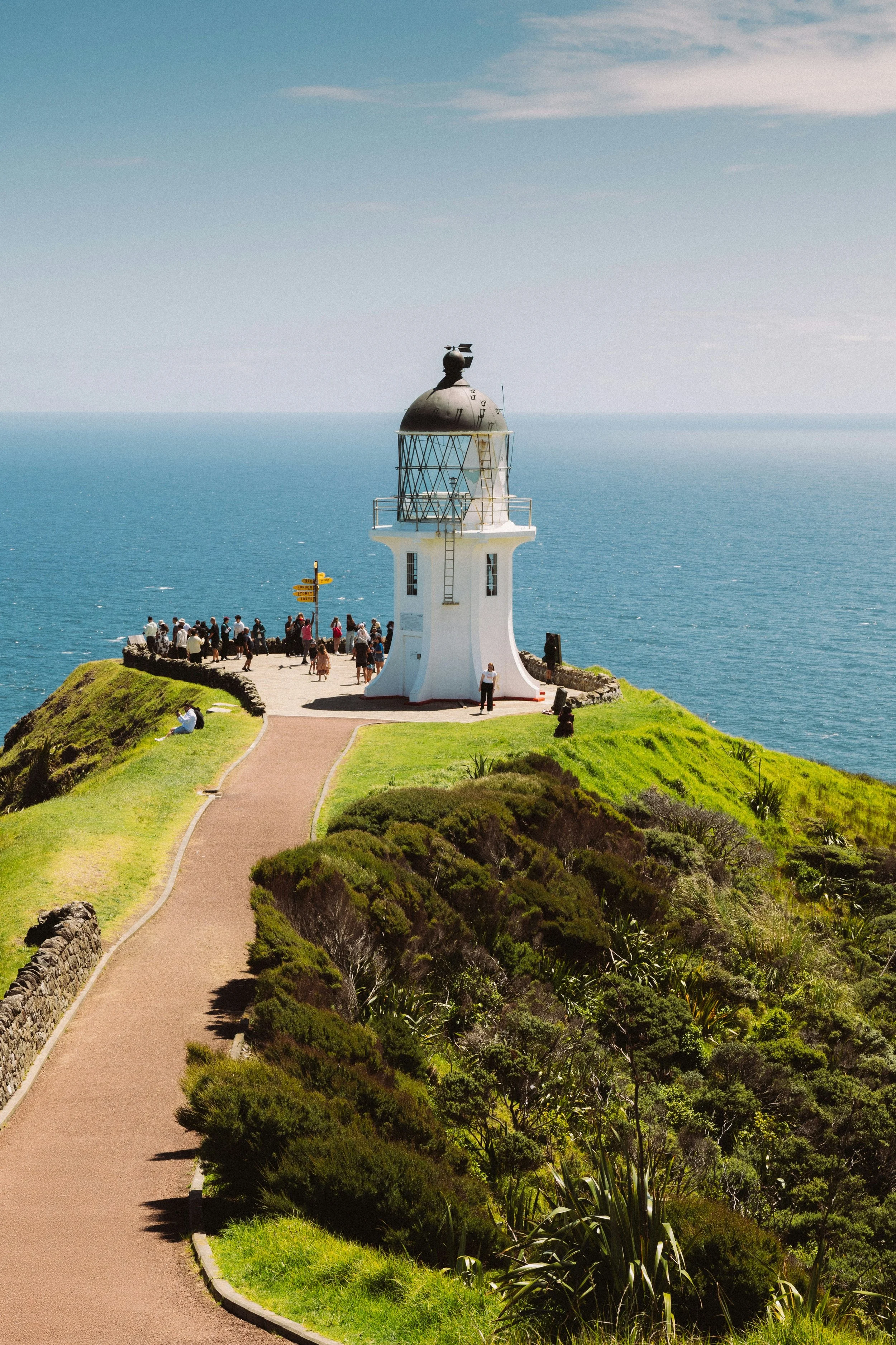 A lighthouse at Cape Te Reinga - Far North, on a grassy hill overlooking the ocean with a group of people nearby, and a clear blue sky.