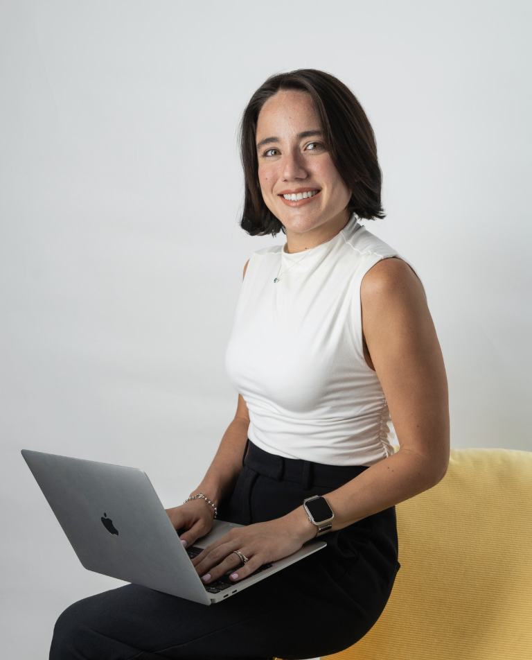Mujer sonriente con cabello castaño oscuro, usando blusa blanca sin mangas, pantalones oscuros, reloj inteligente y anillo, sentada en una silla amarilla con una laptop en las piernas en un fondo gris.
