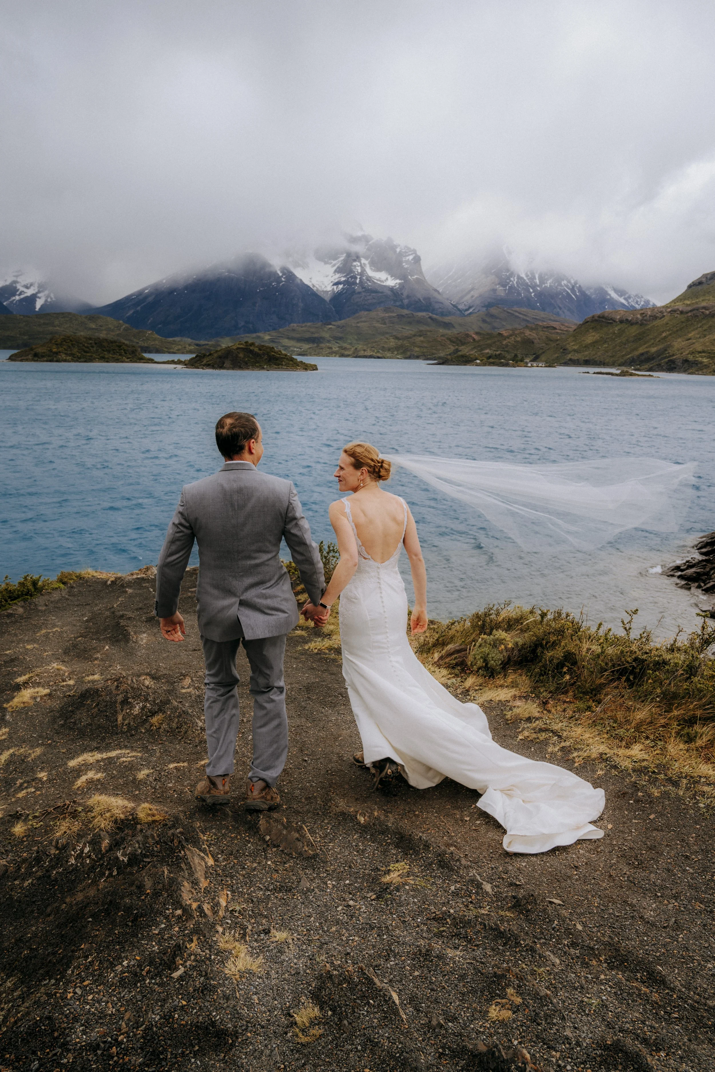 A couple in wedding attire walking hand in hand along a rugged path by a large lake, with snow-capped mountains and cloudy sky in the background.
