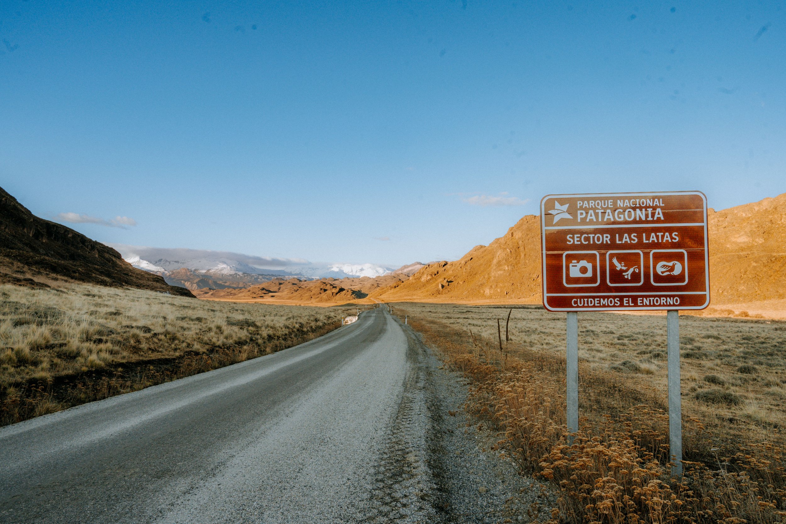 A long dirt road in a mountainous landscape with a sign indicating Parque Nacional Patagonia, Sector Las Latas, cautioning visitors to care for the environment, with a clear blue sky and distant snow-capped mountains.