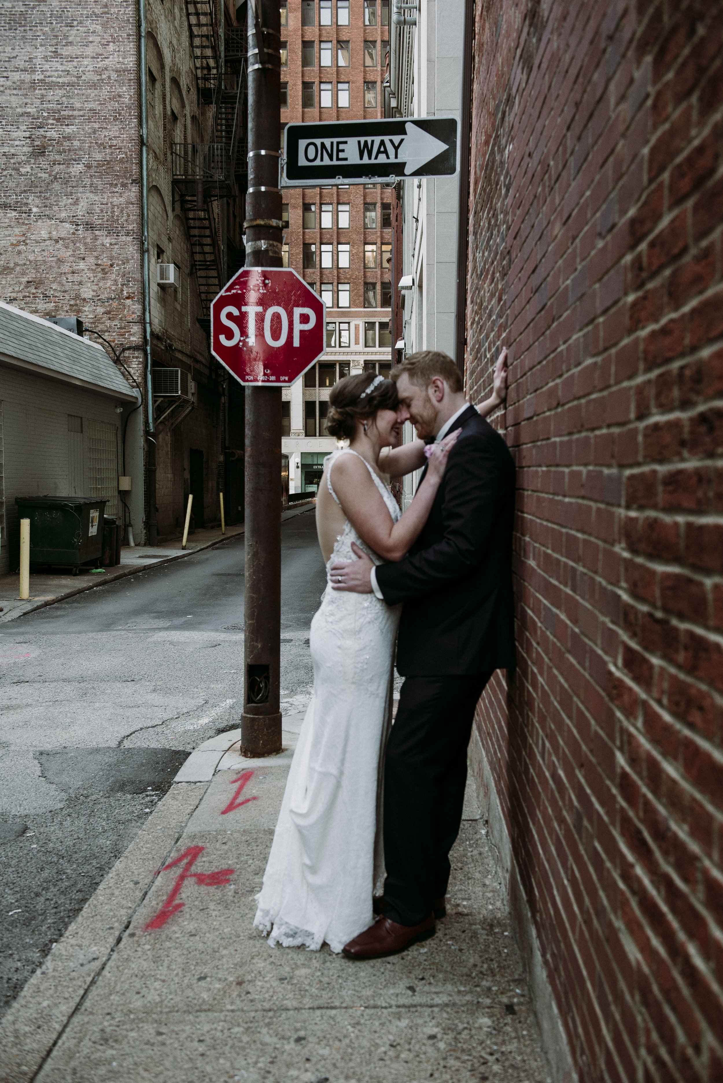 A newlywed couple in wedding attire sharing a tender moment as they stand close against a brick wall in an alleyway with urban buildings in the background. They are smiling and touching foreheads.