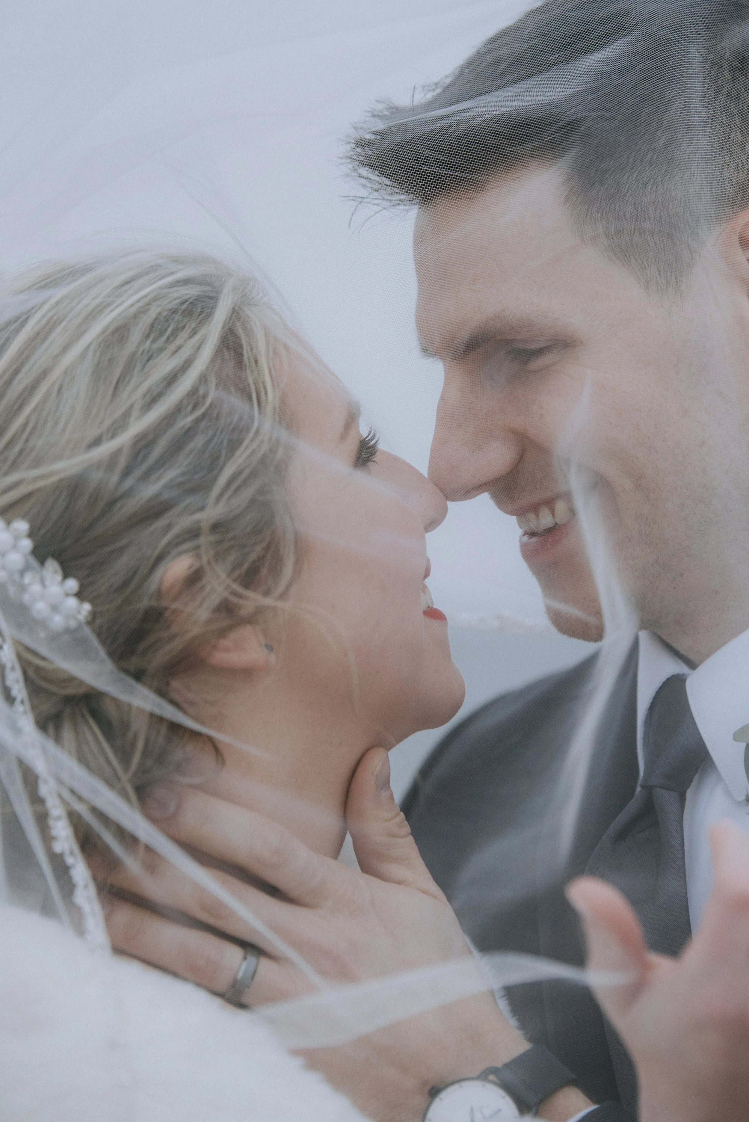 A bride and groom are close together with their foreheads touching, smiling and looking into each other's eyes, under a wedding veil.