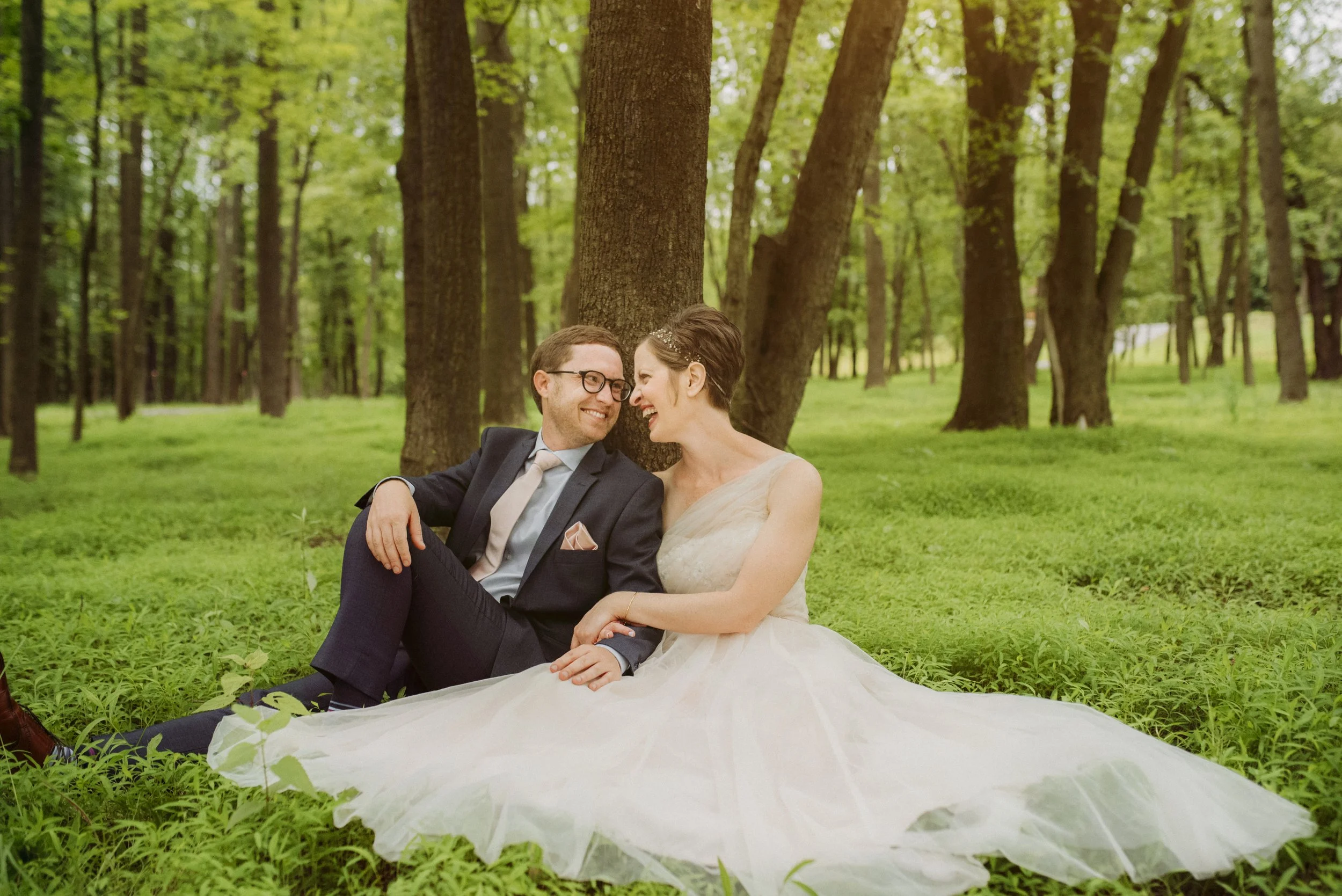 A couple in wedding attire sitting on the grass in a wooded area smiling at each other.