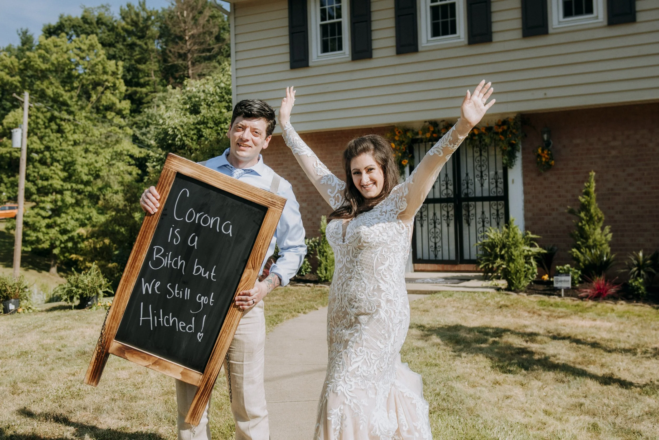 A couple, dressed in wedding attire, standing outside in front of a house with a chalkboard sign that reads, 'Corona is a Bitch but We Still Got Hitch.' The woman is smiling, raising her arm, and wearing a long, elegant lace wedding dress. The man is holding the sign, smiling, and wearing a light-colored shirt with suspenders. There are green trees and a brick house with window shutters in the background.