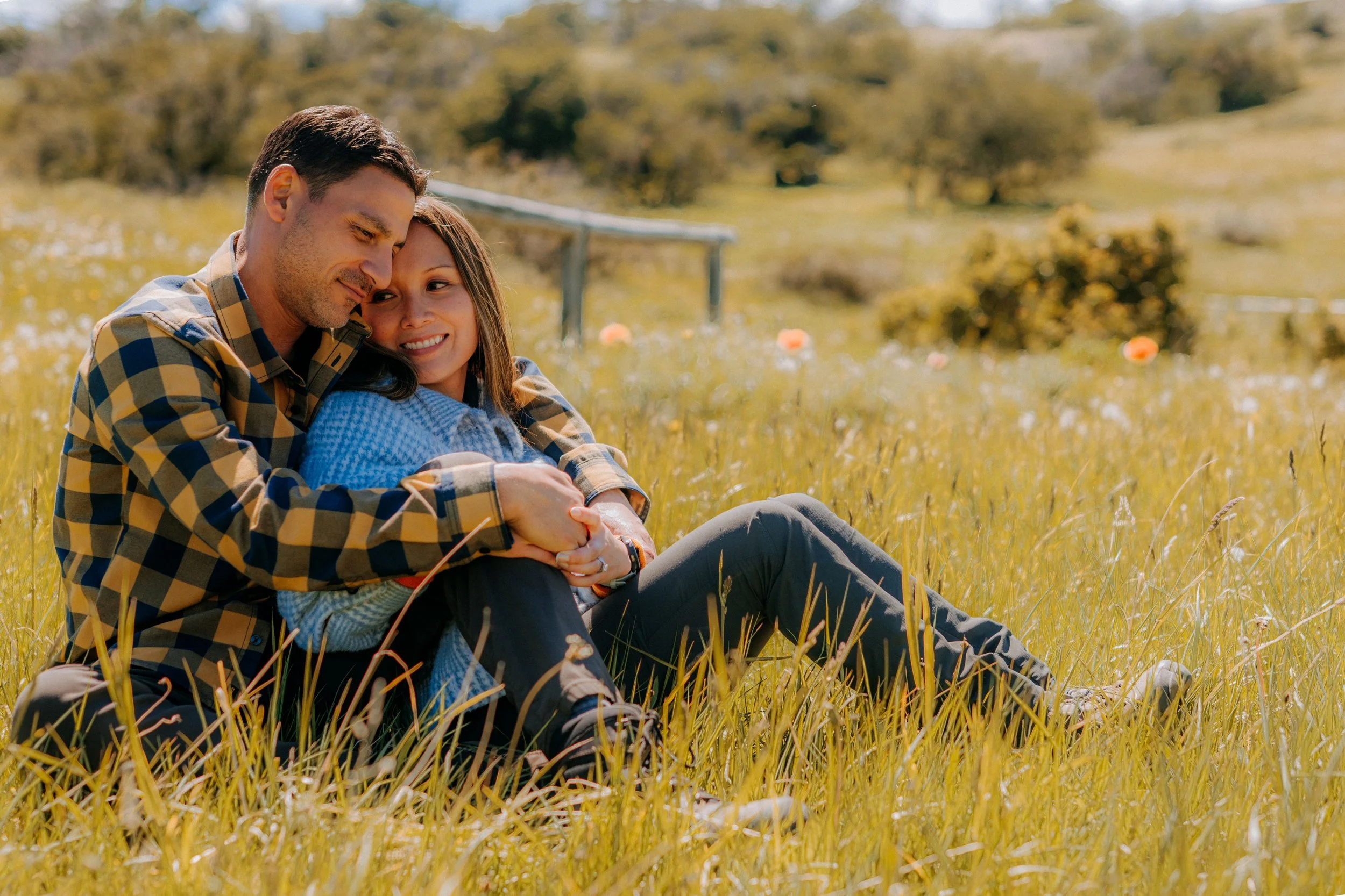 A couple sitting in a grassy field, embracing and smiling, in a natural outdoor setting with trees and a fence in the background.