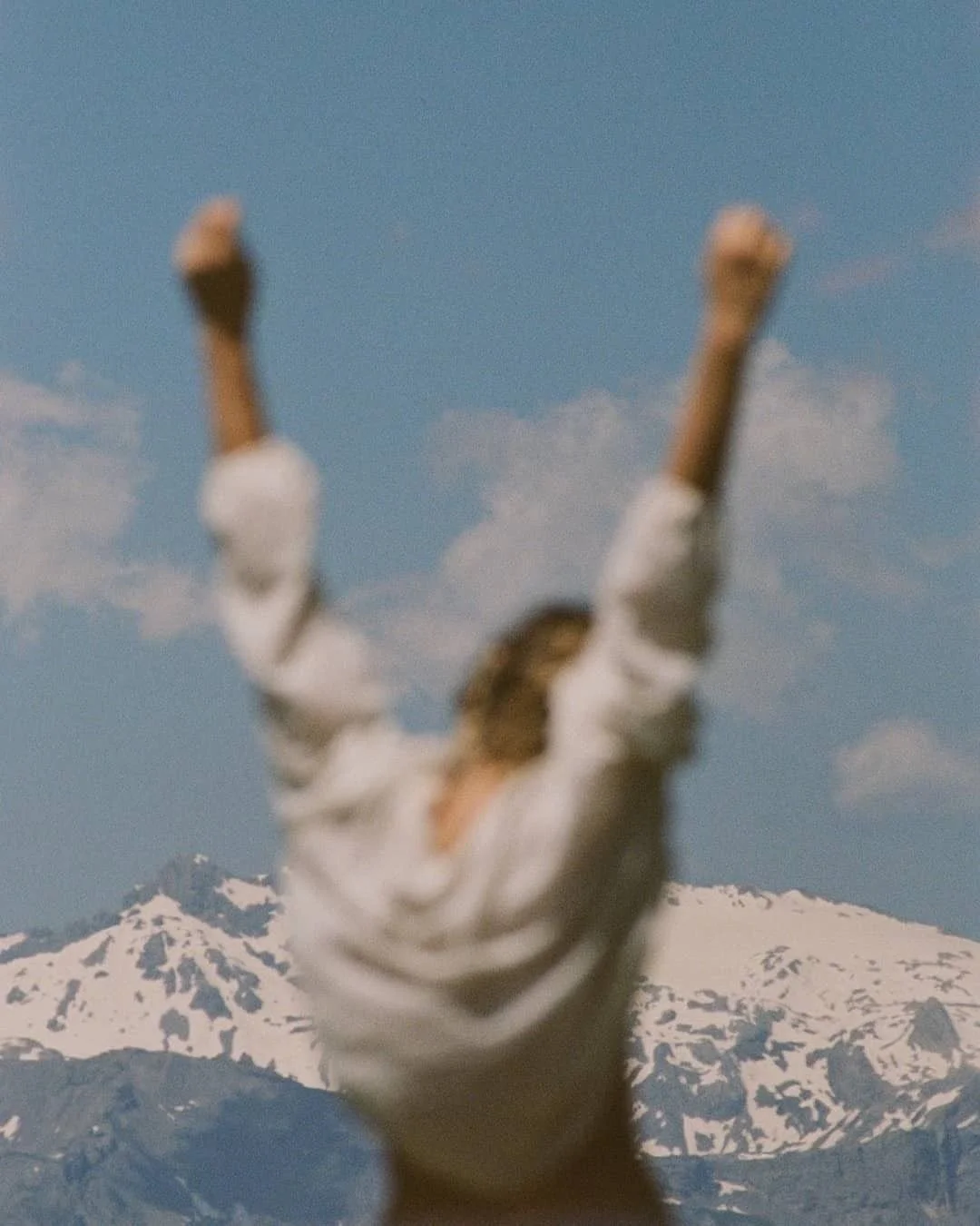 A person in a white shirt with arms raised, standing in front of a snow-capped mountain peak and a blue sky with a few clouds.
