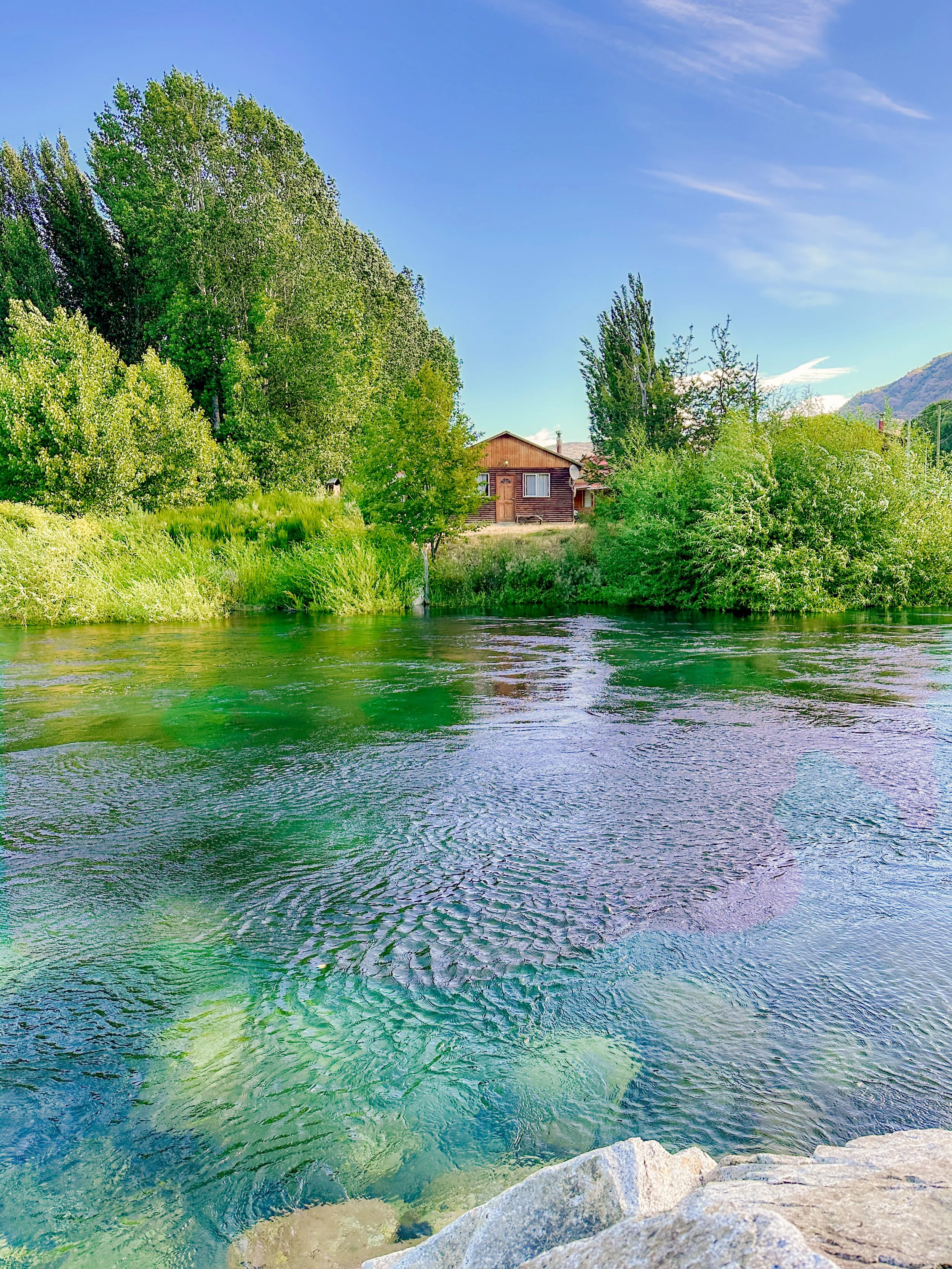 A scenic view of a small wooden house surrounded by green trees and bushes, with a river in the foreground and mountains in the background under a partly cloudy sky.