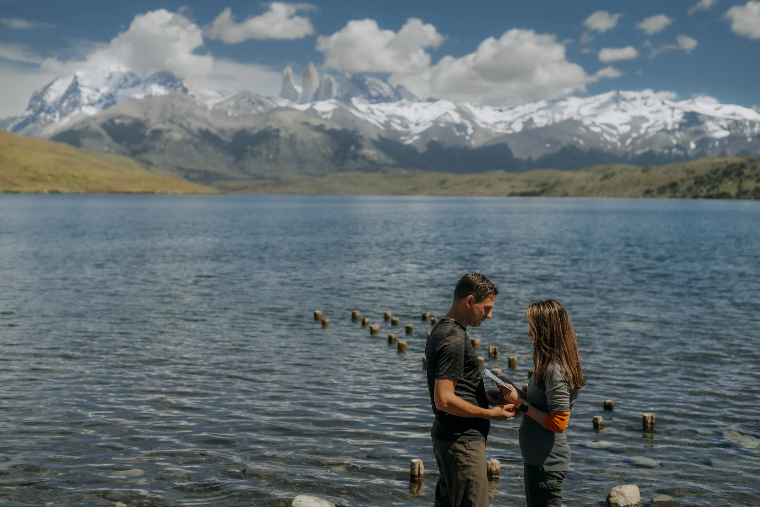 A couple stands close together near a lake with mountains in the background, engaged in a conversation, with the man holding a piece of paper or a book.