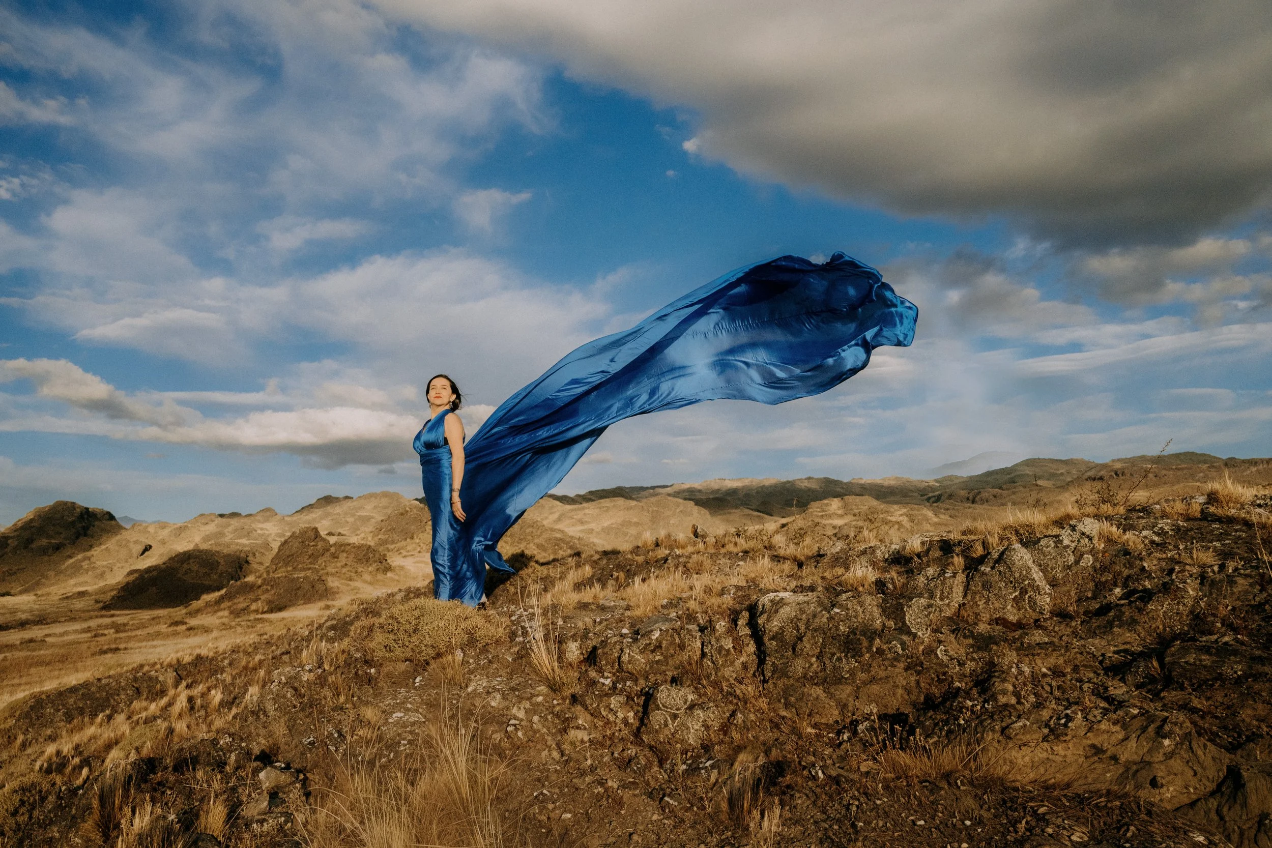 A woman in a blue dress standing on rocky terrain with an expansive sky and clouds behind her, holding a flowing blue fabric that trails in the wind.