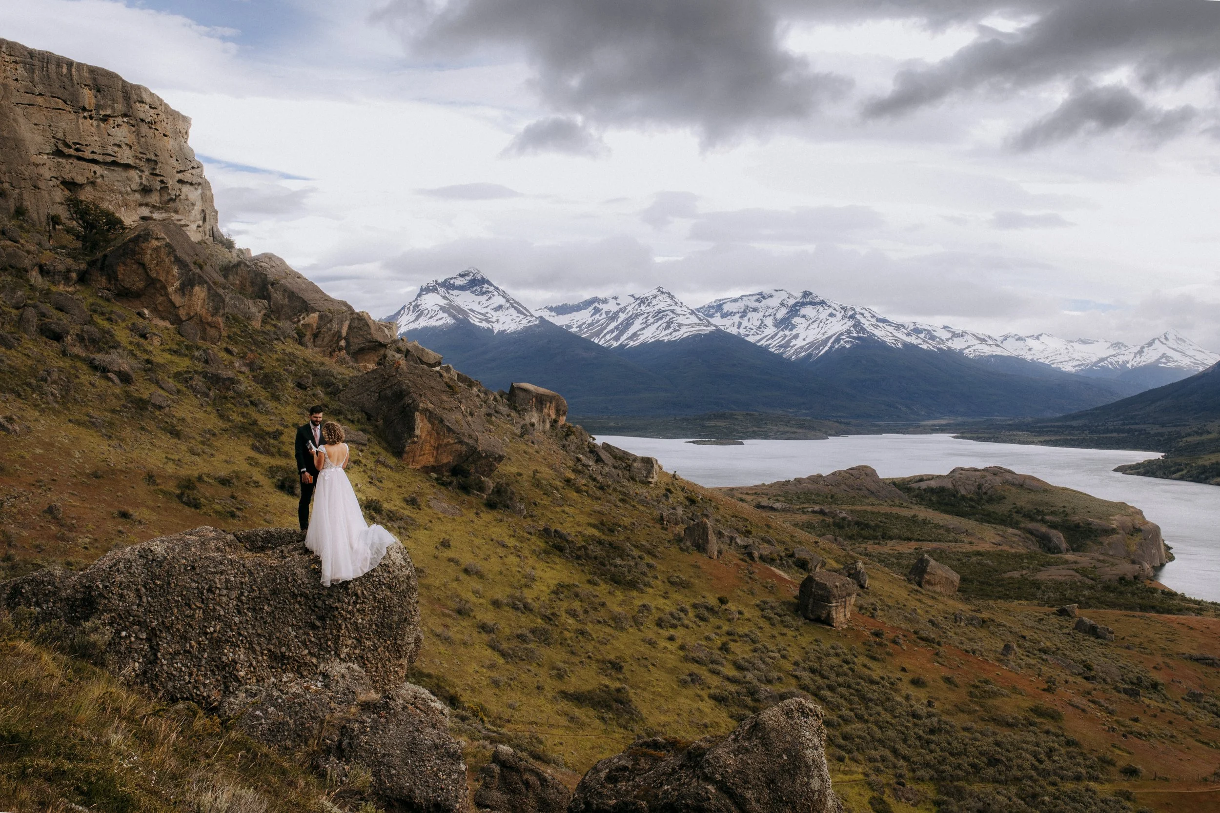 A couple in wedding attire standing on a large rock in a mountainous landscape with snow-capped peaks, a lake, and cloudy sky.