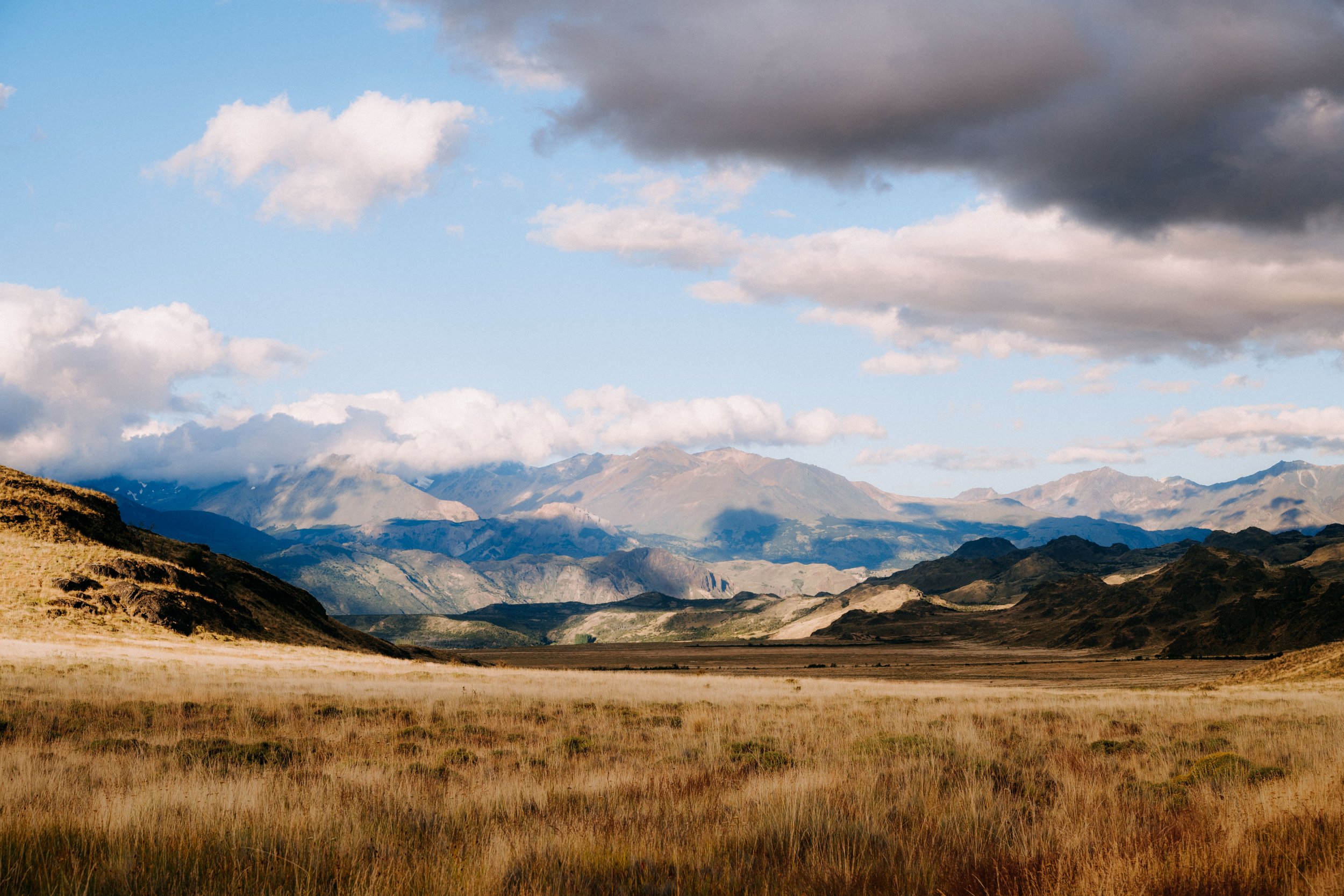Scenic landscape of rolling grassy plains leading to distant mountains under a partly cloudy sky with dark and white clouds.