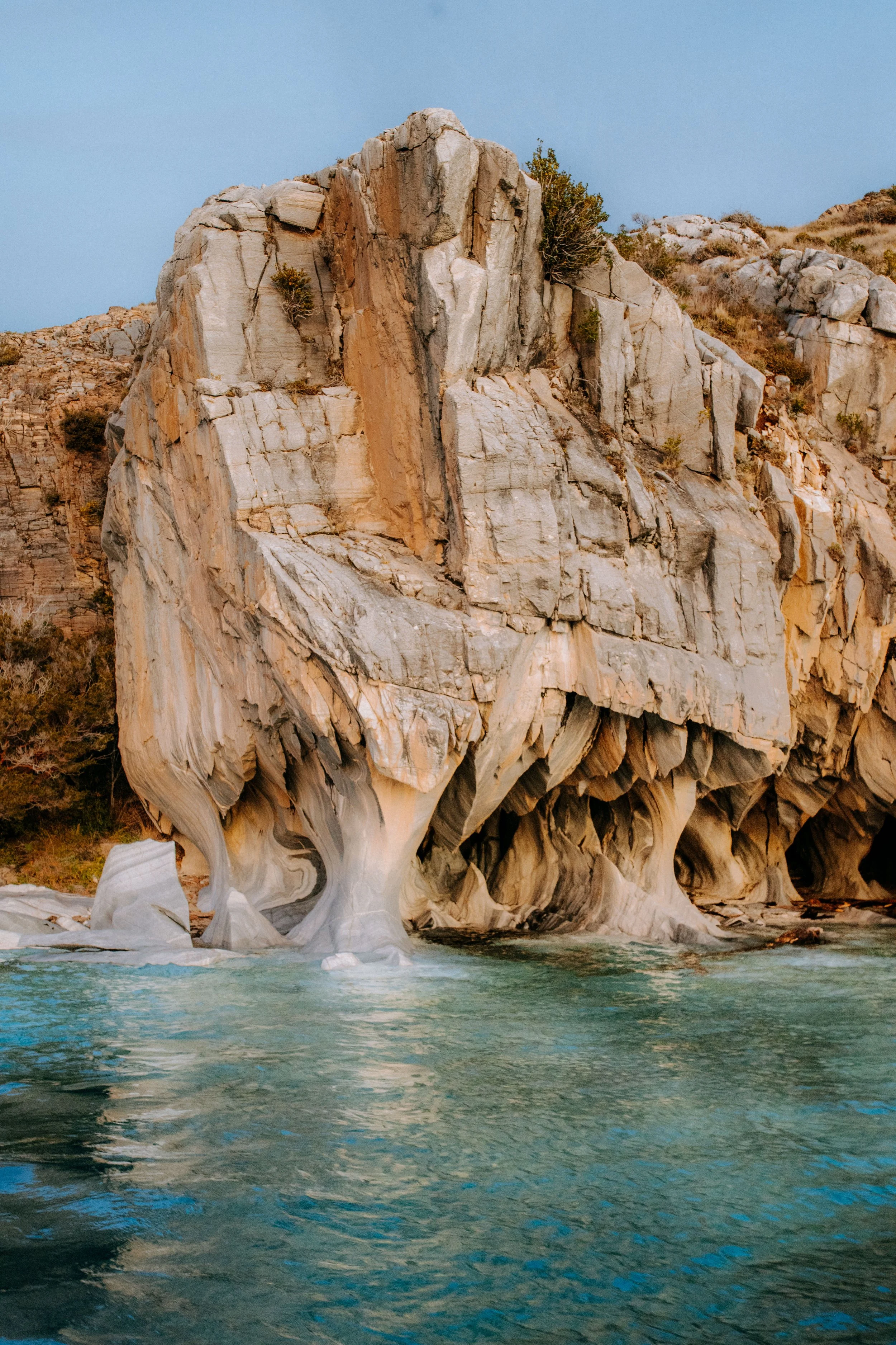A large white and beige marble cliff with layered, textured rock formations rises from the water, with small green shrubs growing in its crevices. The cliff's base is smooth, curving into calm, turquoise water, and a clear sky is above.