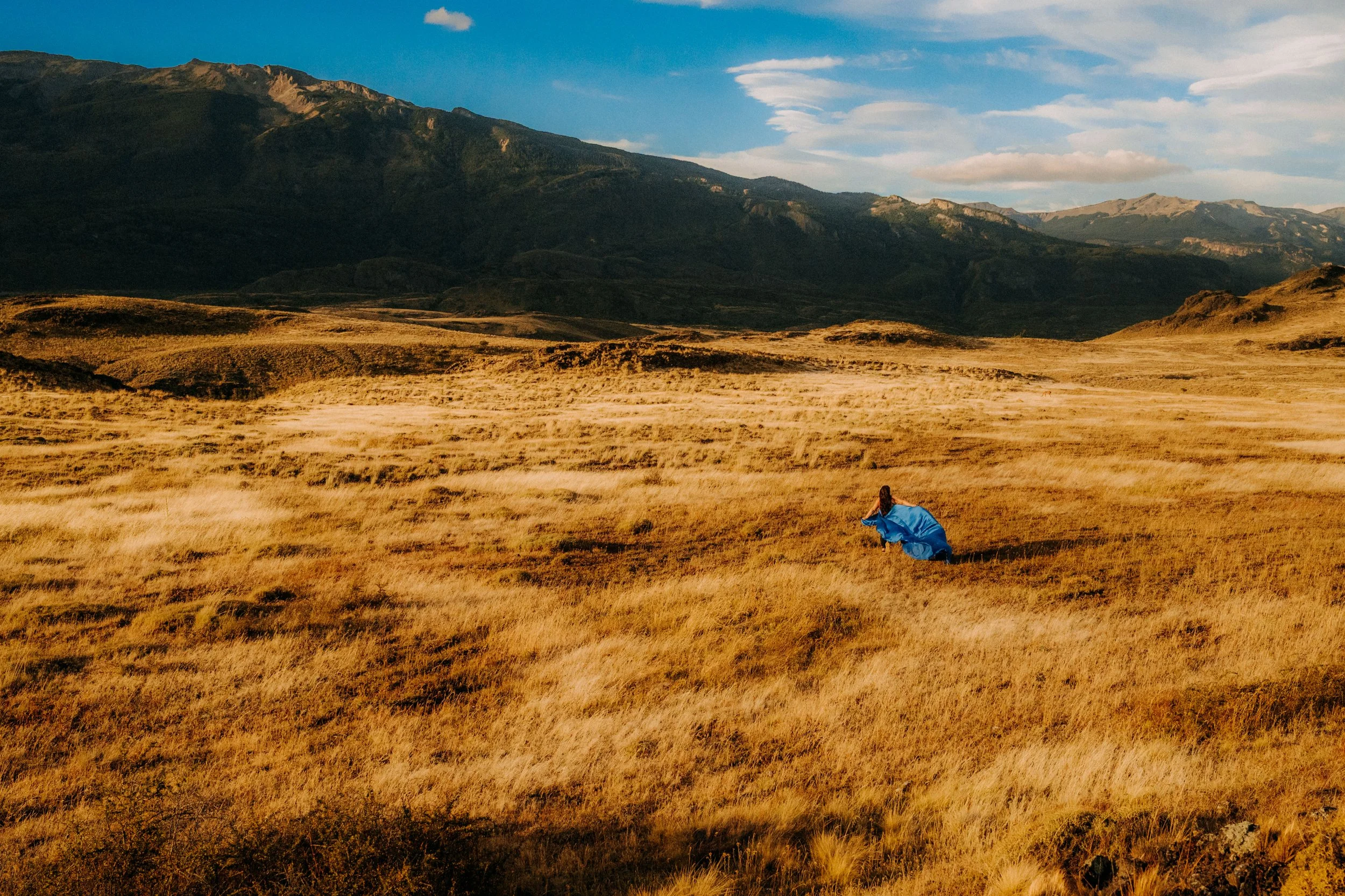 A person in a blue jacket is sitting or lying on a grassy hillside with golden grass. The background features mountains and a partly cloudy sky.