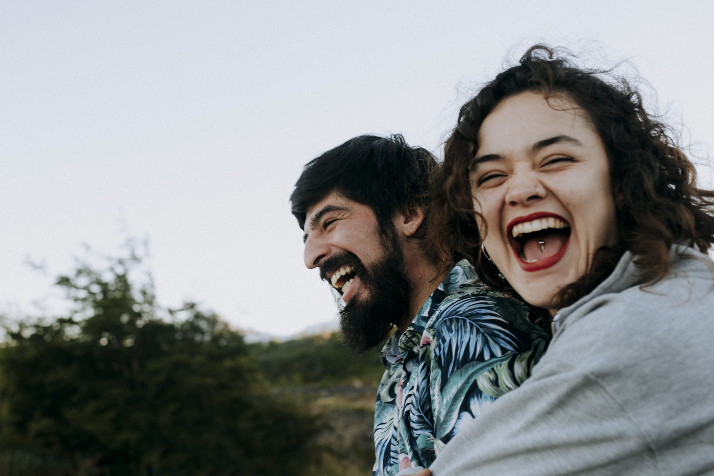 Two people laughing outdoors, one with dark hair and beard, the other with curly brown hair, against a cloudy sky and trees in the background.