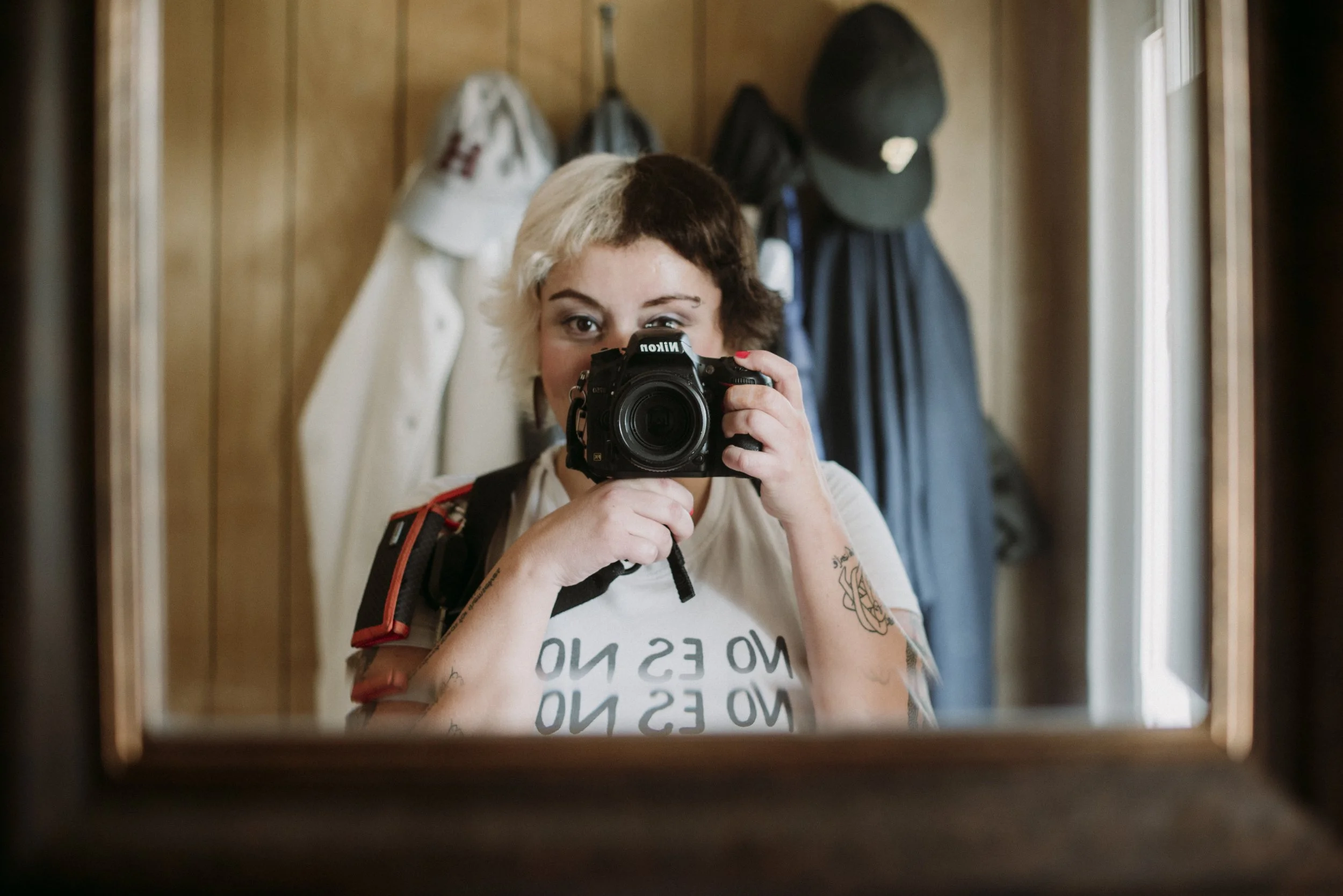 Person taking a photograph of themselves in a mirror, showing partly a face with short hair and tattoos on the arm, with a background of hanging coats and backpacks.