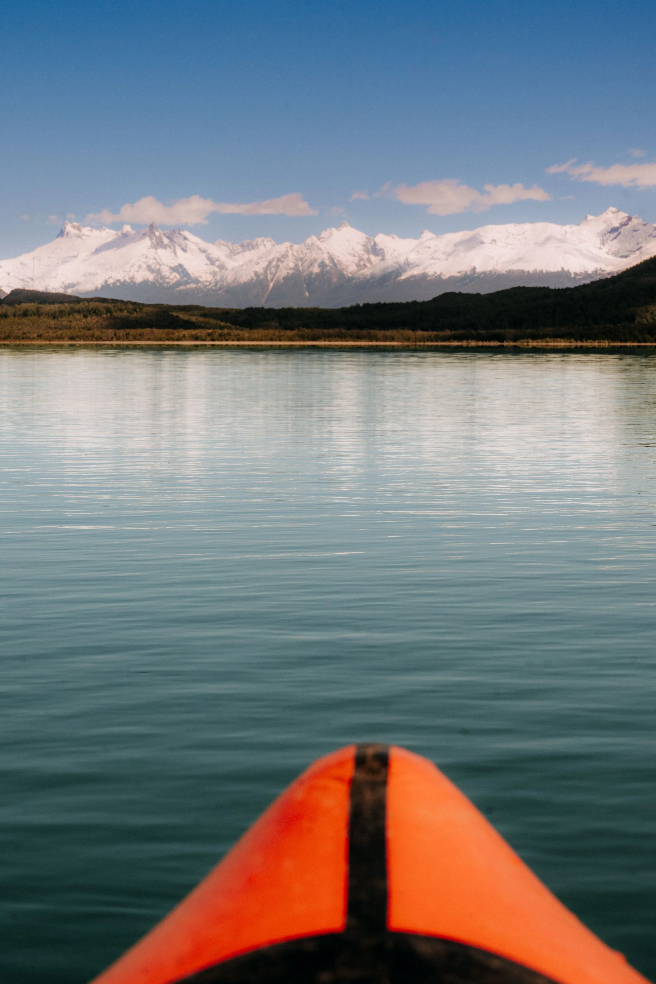 View from a kayak on calm water facing snow-capped mountains under a blue sky.