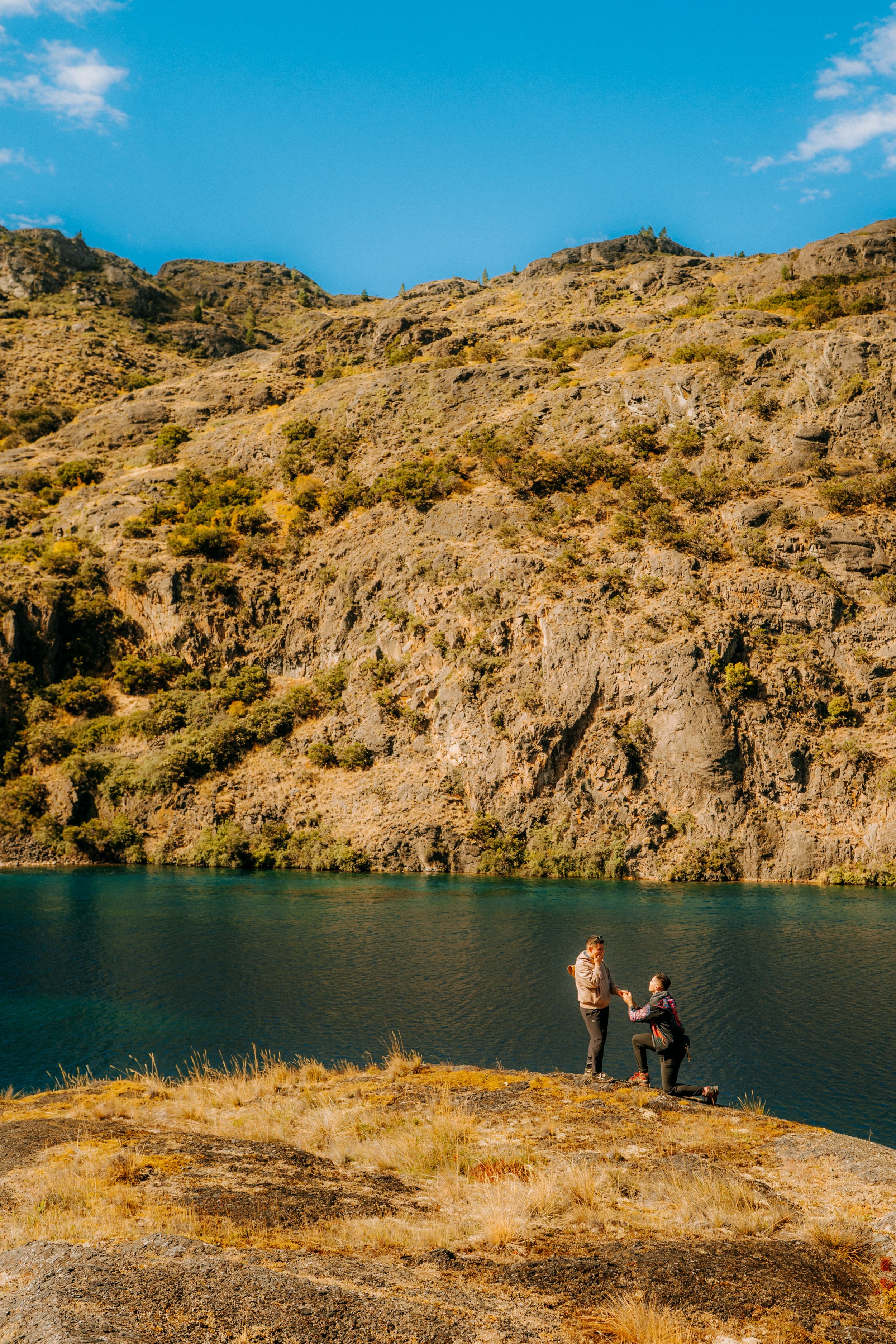 A man kneeling on grassy ground, proposing to a woman near a lake with a hillside covered in rocks and dry bushes in the background, under a blue sky with a few clouds.