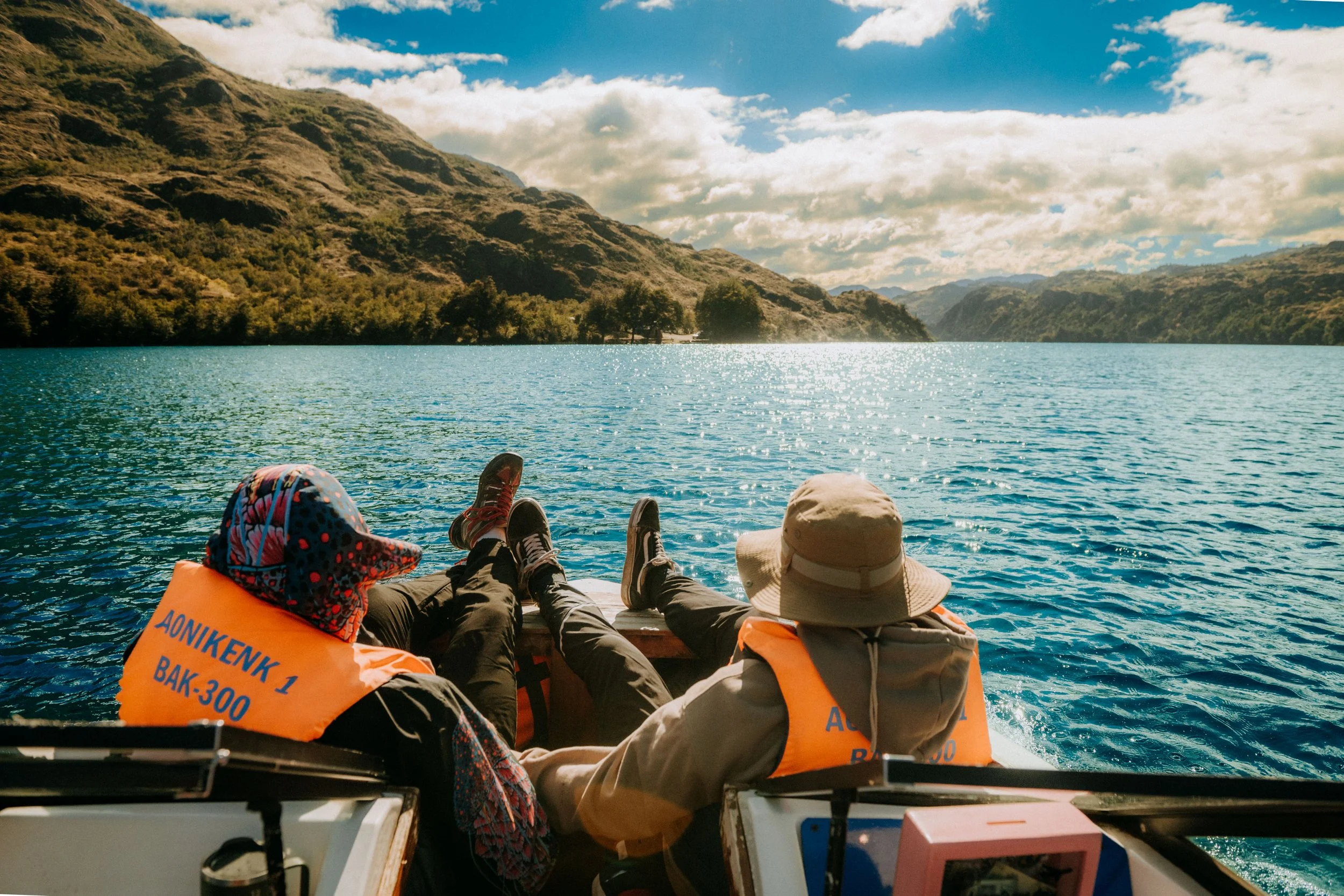 Three people lying on a boat, wearing bright orange life jackets, relaxing and looking at a lake with mountains in the background under a partly cloudy sky.