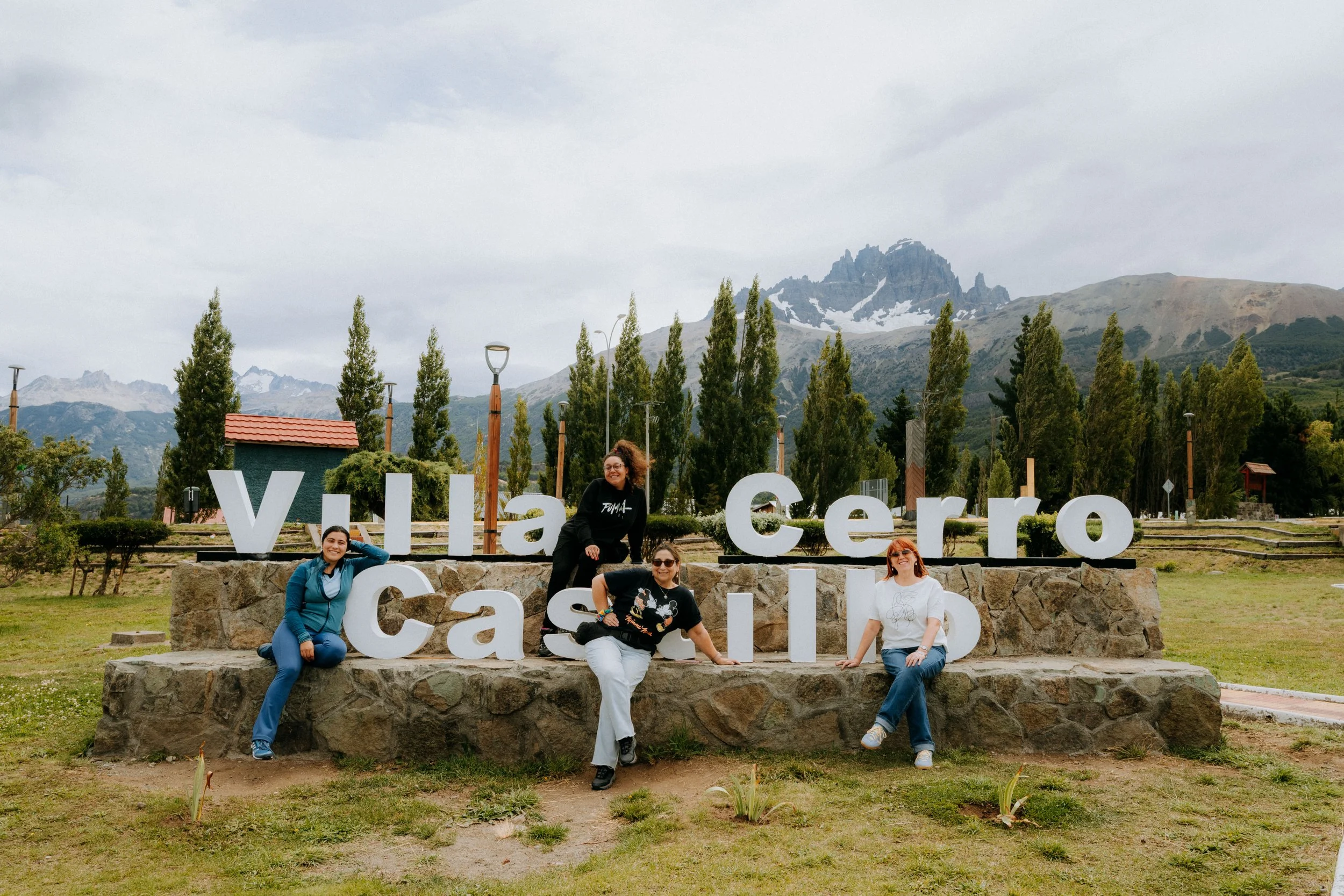 A group of five women posing in front of a large sign that reads 'Yllasero Casapup' with a mountainous landscape in the background.