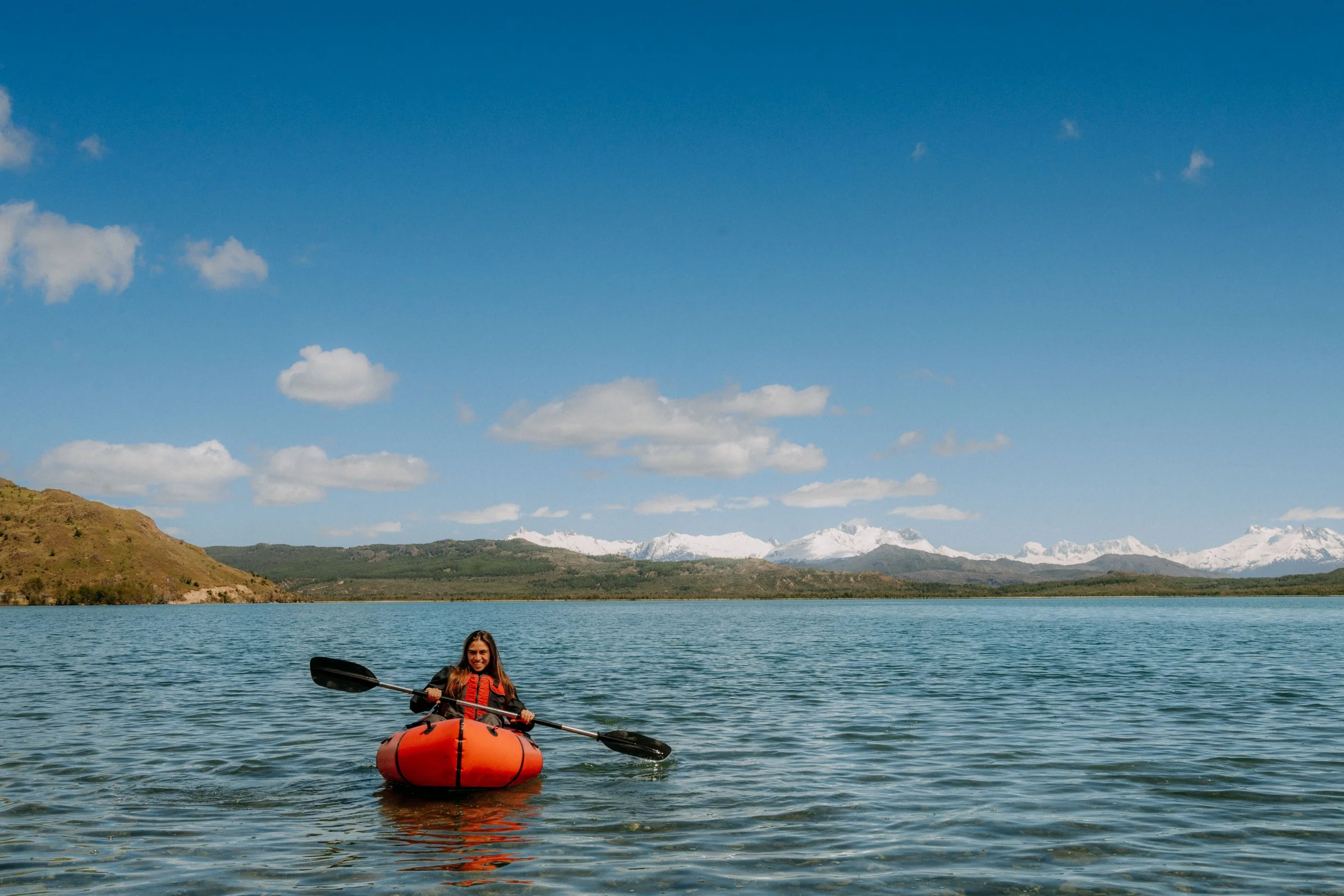 A woman kayaking on a lake with mountains and a blue sky with clouds in the background.
